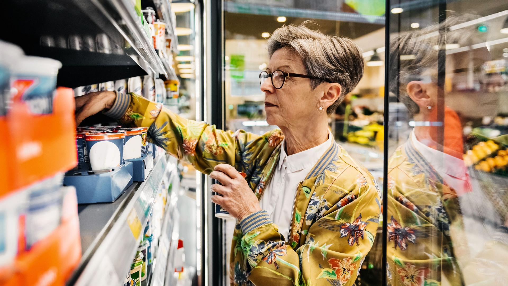 Eine ältere Frau im Supermarkt, die Lebensmittel aus einem Kühlschrank herausholt.