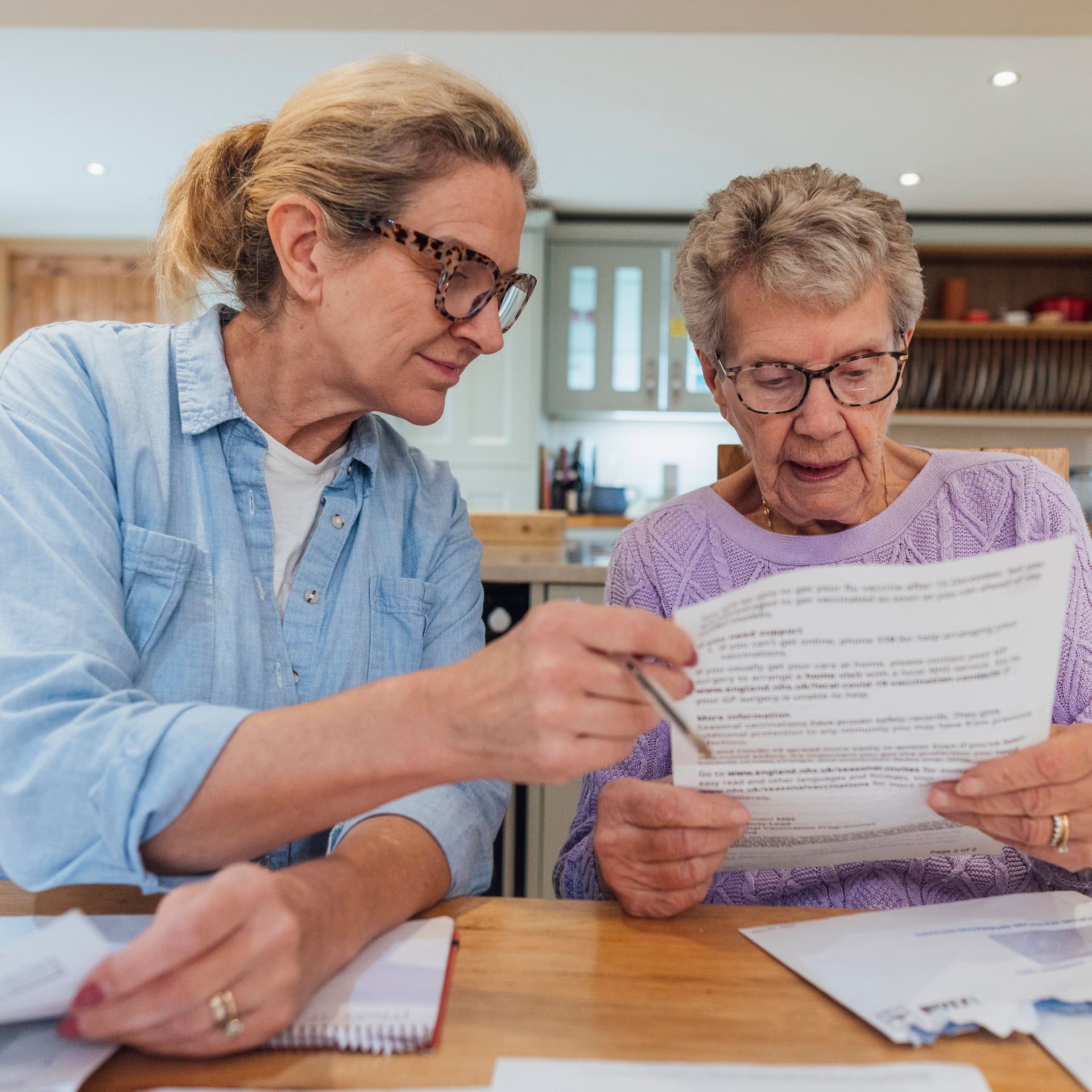 Eine ältere Frau und ihre erwachsene Tochter sitzen am Esstisch und schauen besorgt Finanzrechnungen durch.
