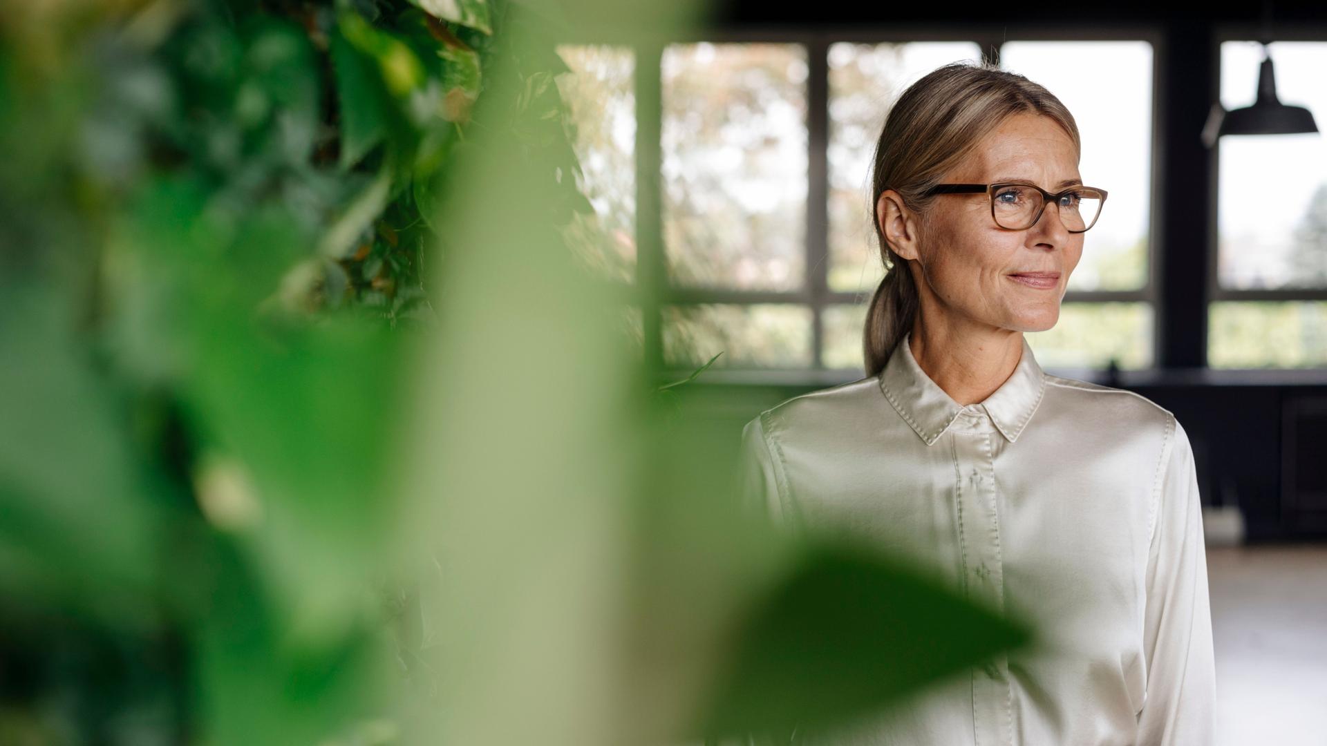 Lächelnde mittelalte Frau mit Brille und schicker Bluse im Büro guckt zur Seite. Im Vordergrund sind grüne Pflanzen.