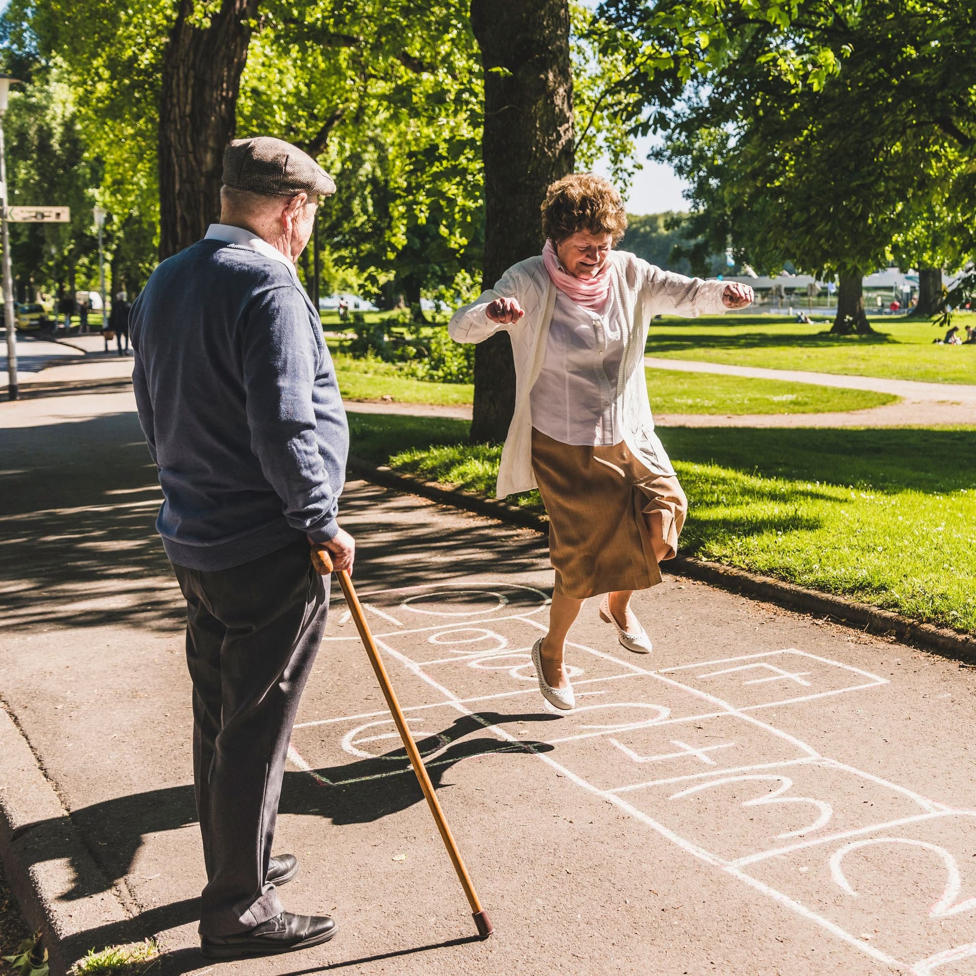 Seniorin springt in einem Stadtpark durch ein Himmel und Hölle Spiel, ein Mann mit Gehstock sieht ihr dabei zu.