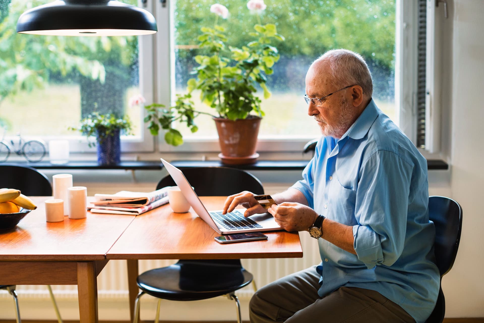 Älterer Mann sitzt mit einem Laptop an einem Tisch und hat eine Kreditkarte in der Hand.