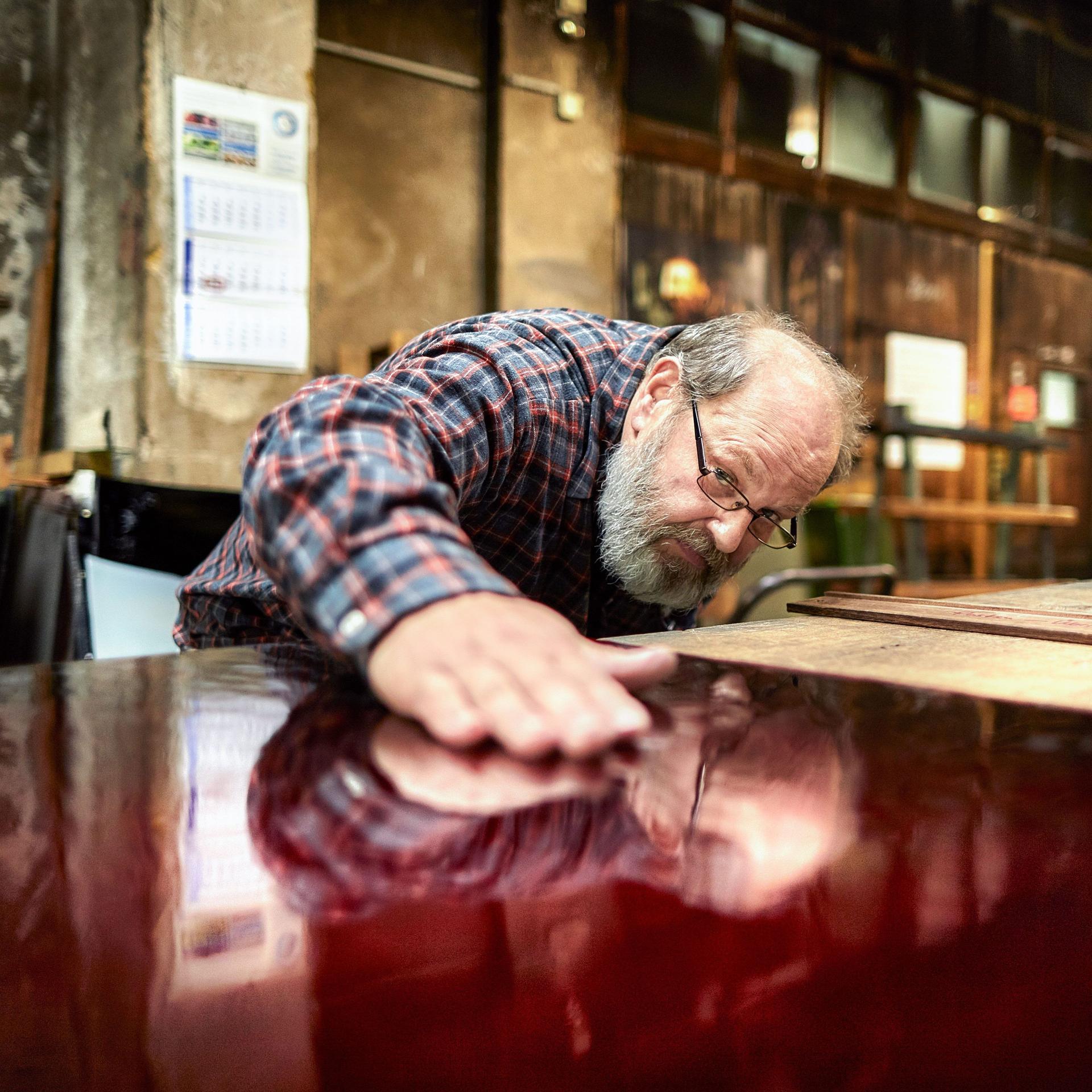 Ein älterer Mann mit Brille und Vollbart beugt sich in einer Werkstatt über eine glatte, rot lackierte Oberfläche und hält die flache Hand darauf.