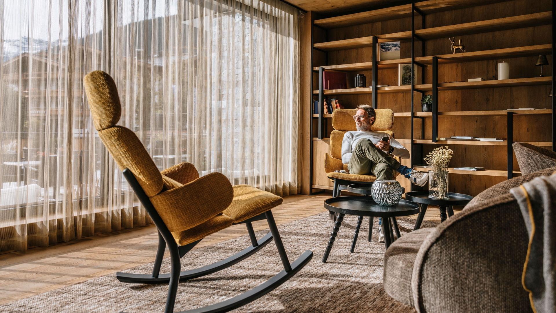 Mann mit Smartphone sitzt in einem Wohnzimmer mit Teppichboden und Blick auf Holzhäuser und Berge.