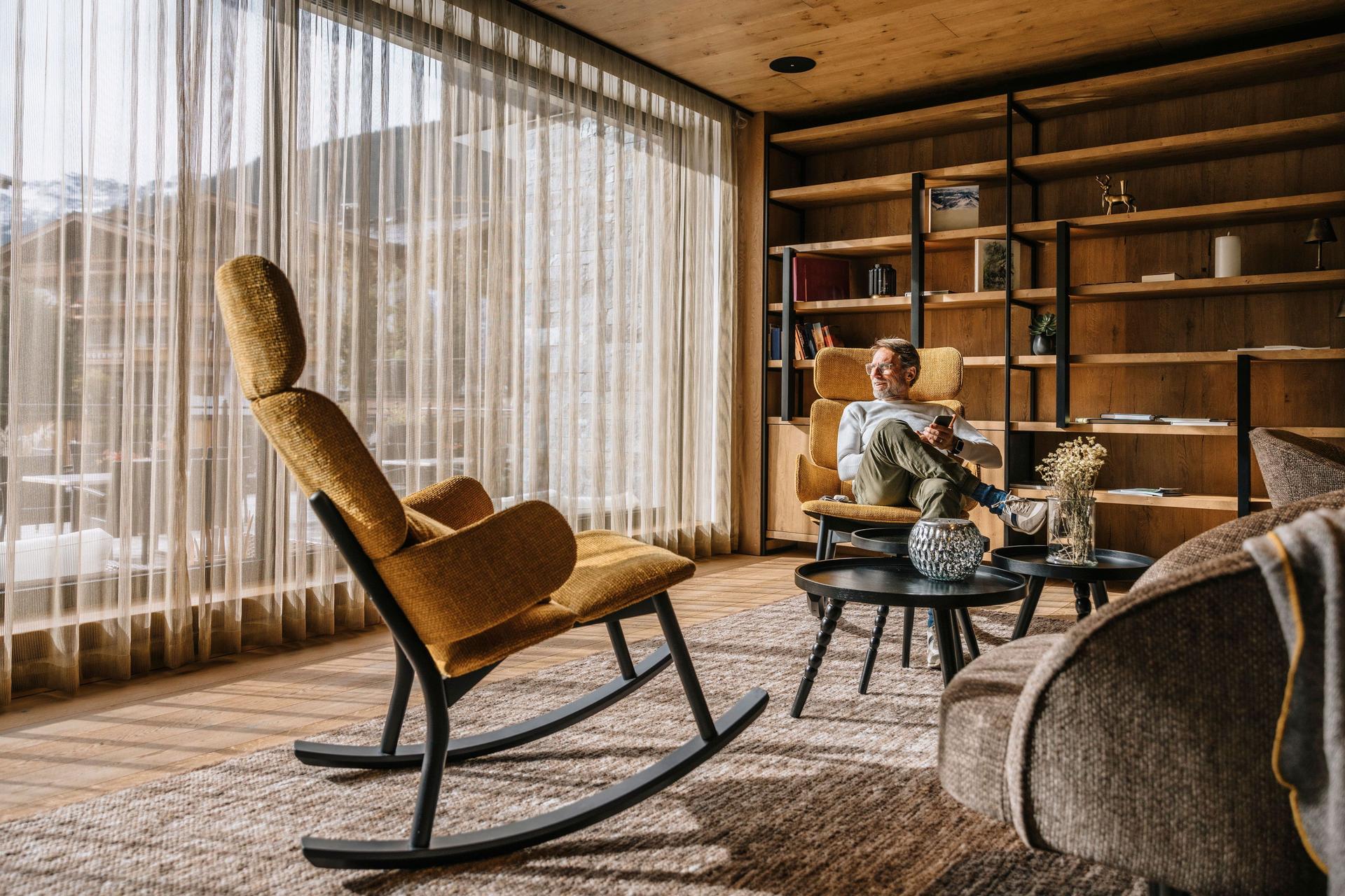 Mann mit Smartphone sitzt in einem Wohnzimmer mit Teppichboden und Blick auf Holzhäuser und Berge.