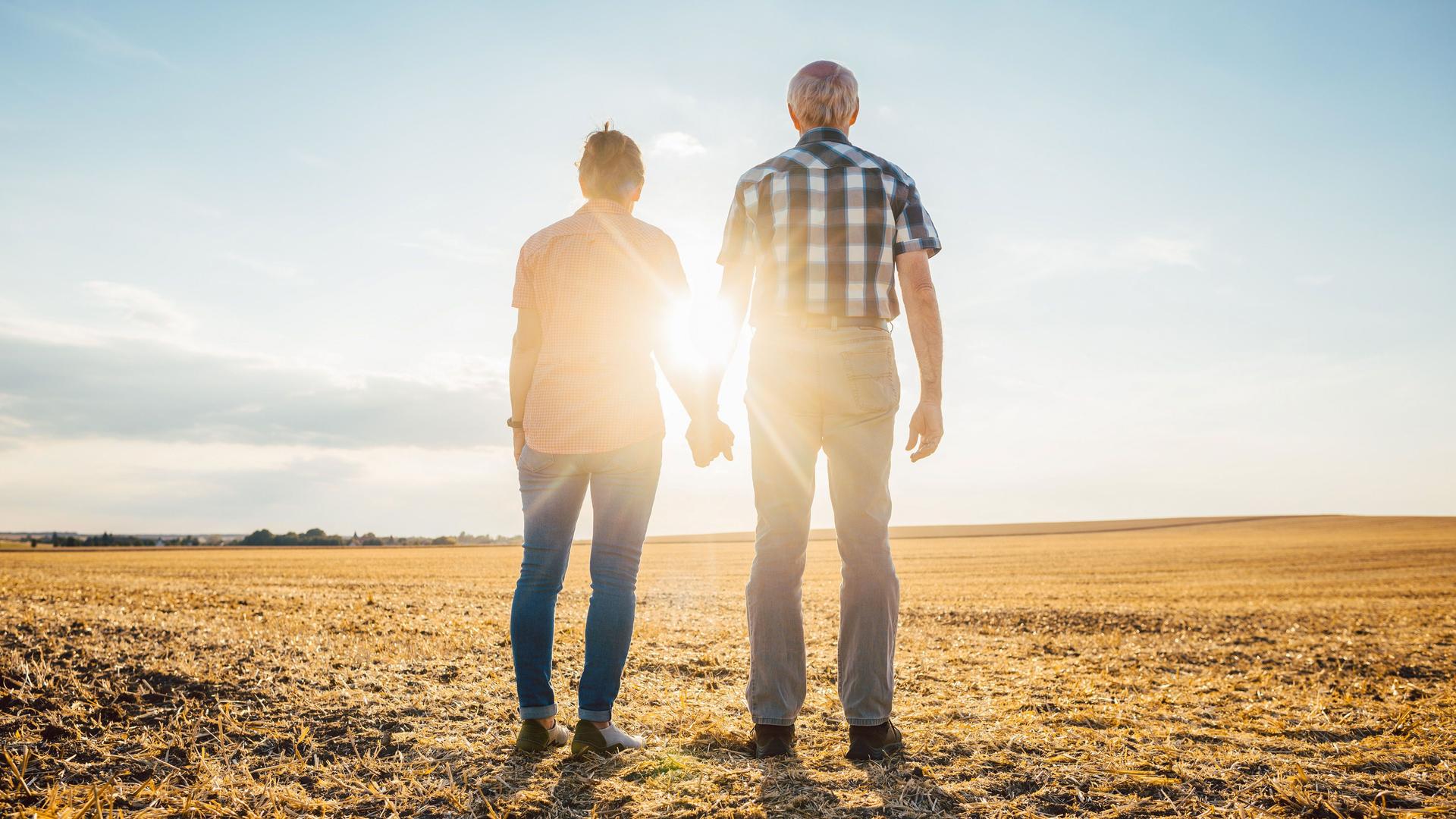 Älteres Paar geht Hand in Hand auf einem Feld im Sonnenuntergang spazieren.
