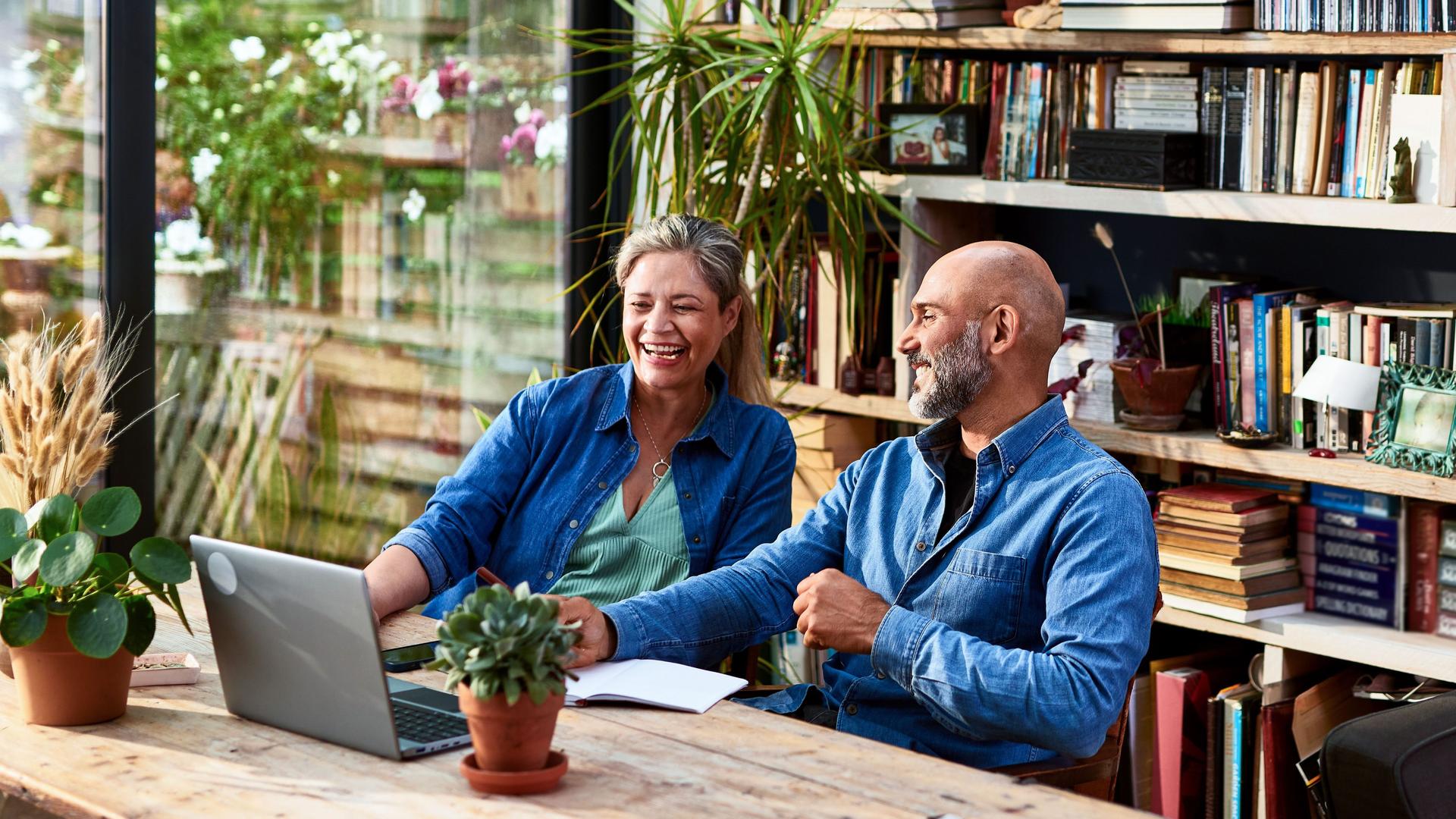 Älteres Paar, das zusammen an einem Holztisch vor einem Laptop sitzt und miteinander redet und lacht. Hinter ihnen ist ein Bücherregal und ein Panoramafenster in den Garten.