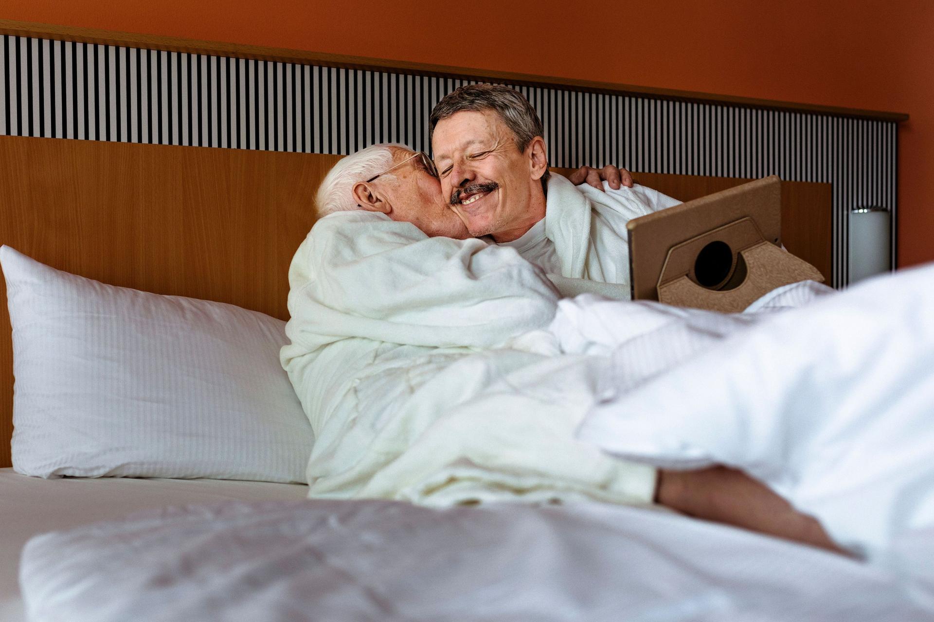 Zwei reifere Männer, die in Bademänteln in einem Bett liegen und sich umarmen. Ein Mann küsst den anderen auf die Backe während dieser ein Tablet auf dem Schoß hat.