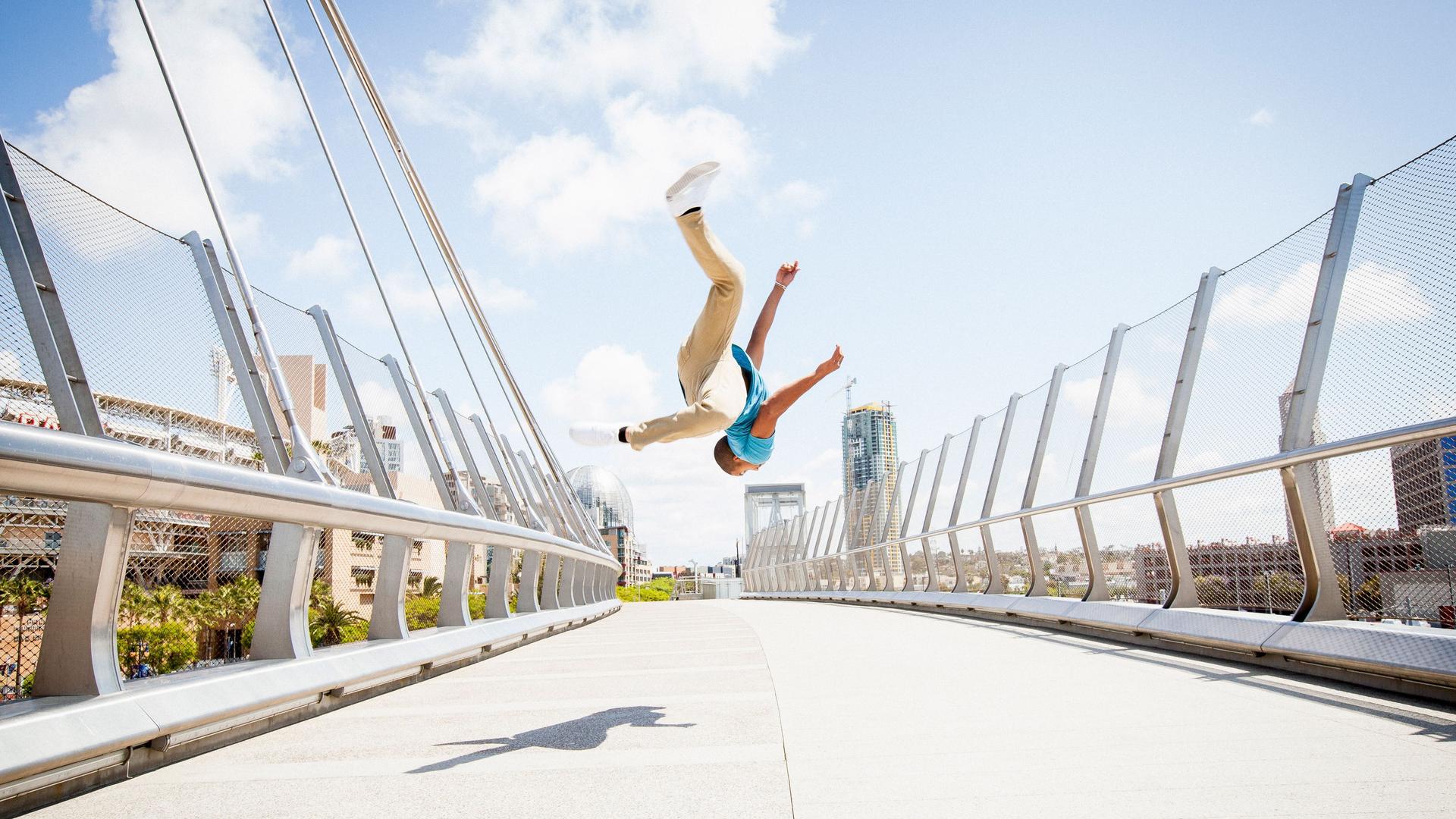 Junger Mann, der auf einer Fußgängerbrücke einen Salto schlägt. Er schwebt vor einem blauen Himmel in der Luft.
