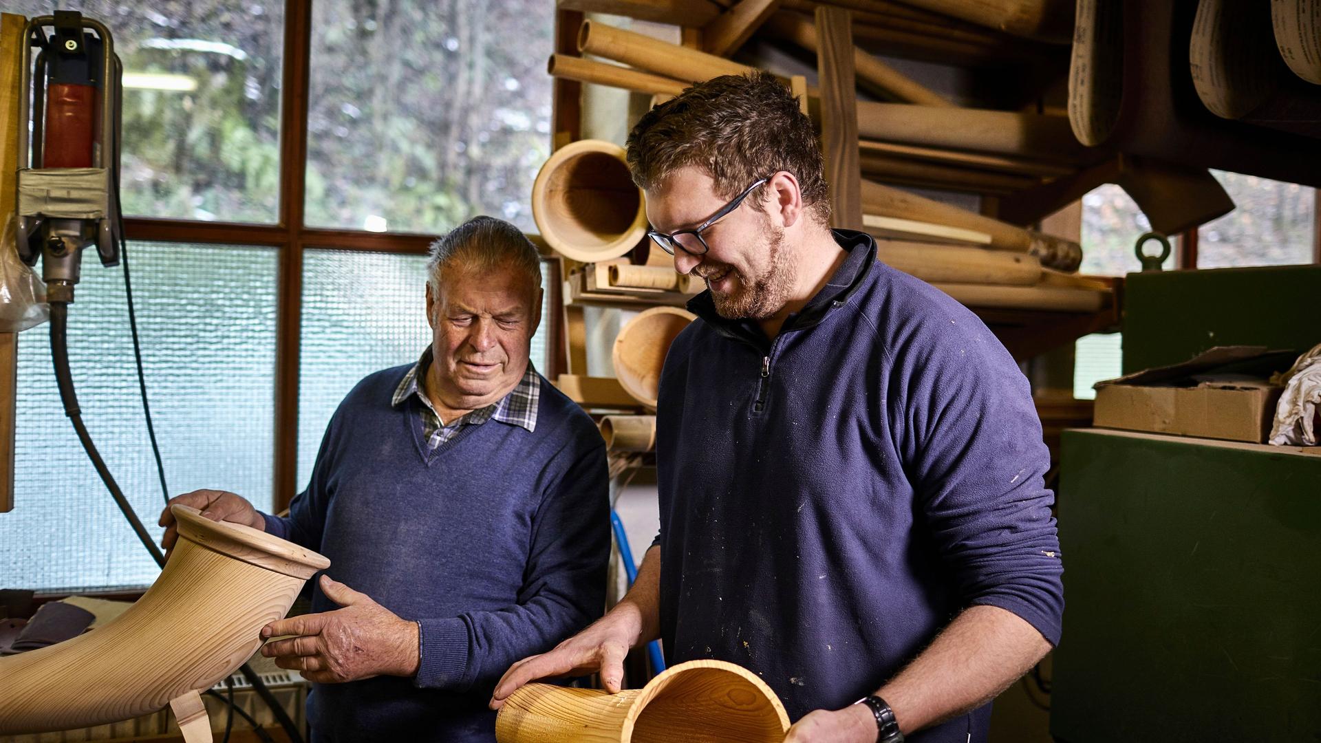 Alter und junger Instrumentenbauer in einer Werkstatt arbeiten an Alphörnern und lächeln dabei.