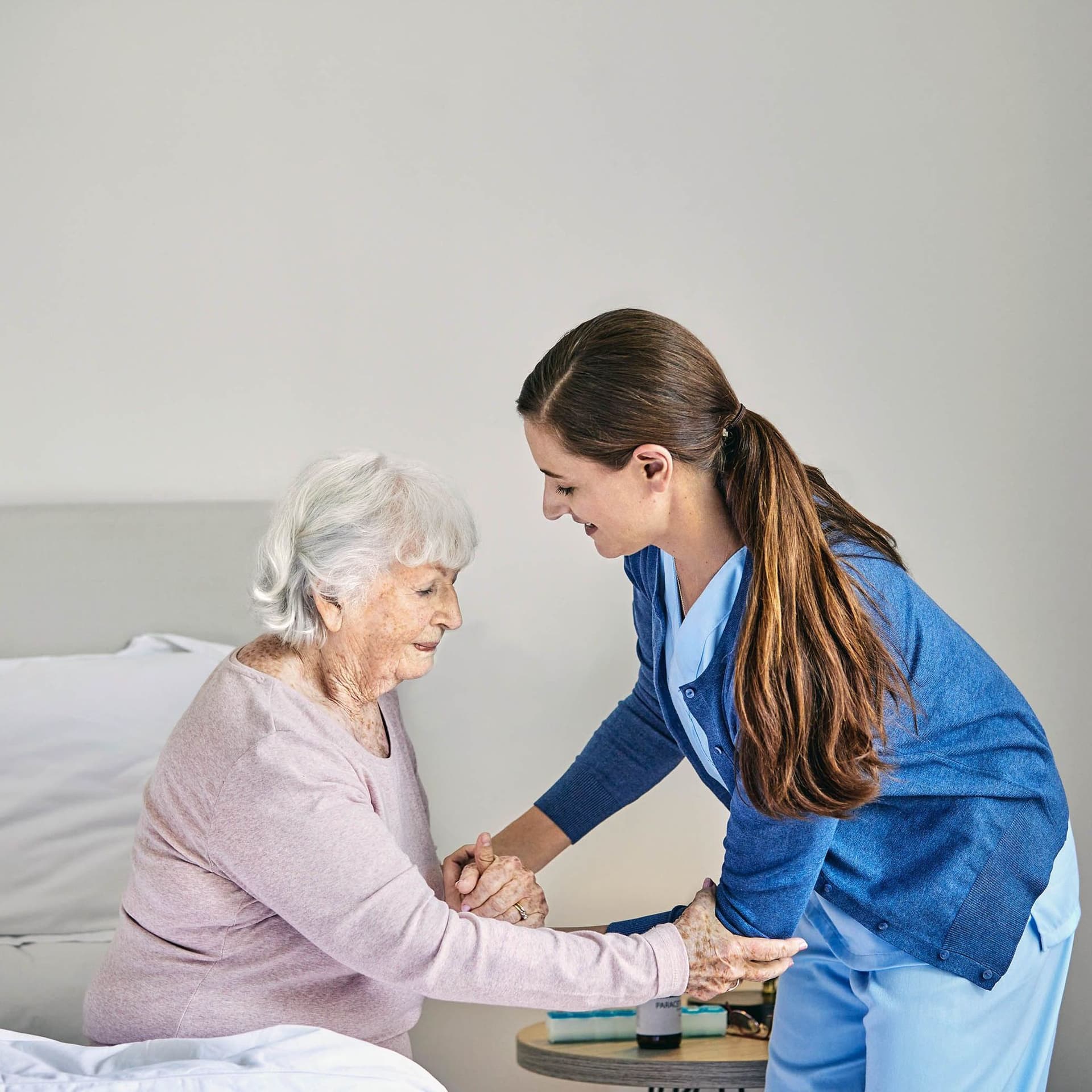 Eine junge Pflegerin hilft einer älteren Frau, die am Bettrand sitzt, beim Aufstehen. Sie hält die Seniorin unterstützend an den Armen. Der Raum ist hell und schlicht eingerichtet.