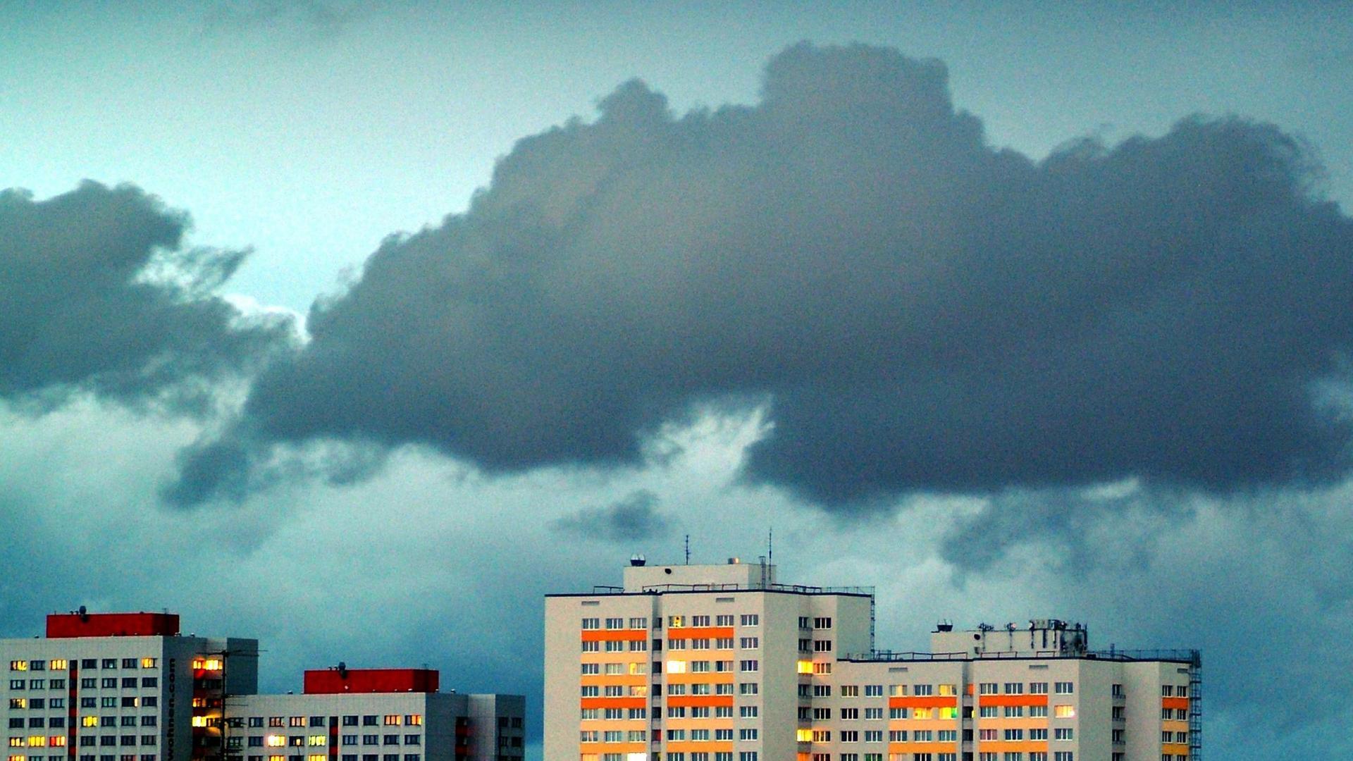 Dunkle Wolken in einem weiten Himmel ziehen über Hochhäuser deren Fenster beleuchtet sind.