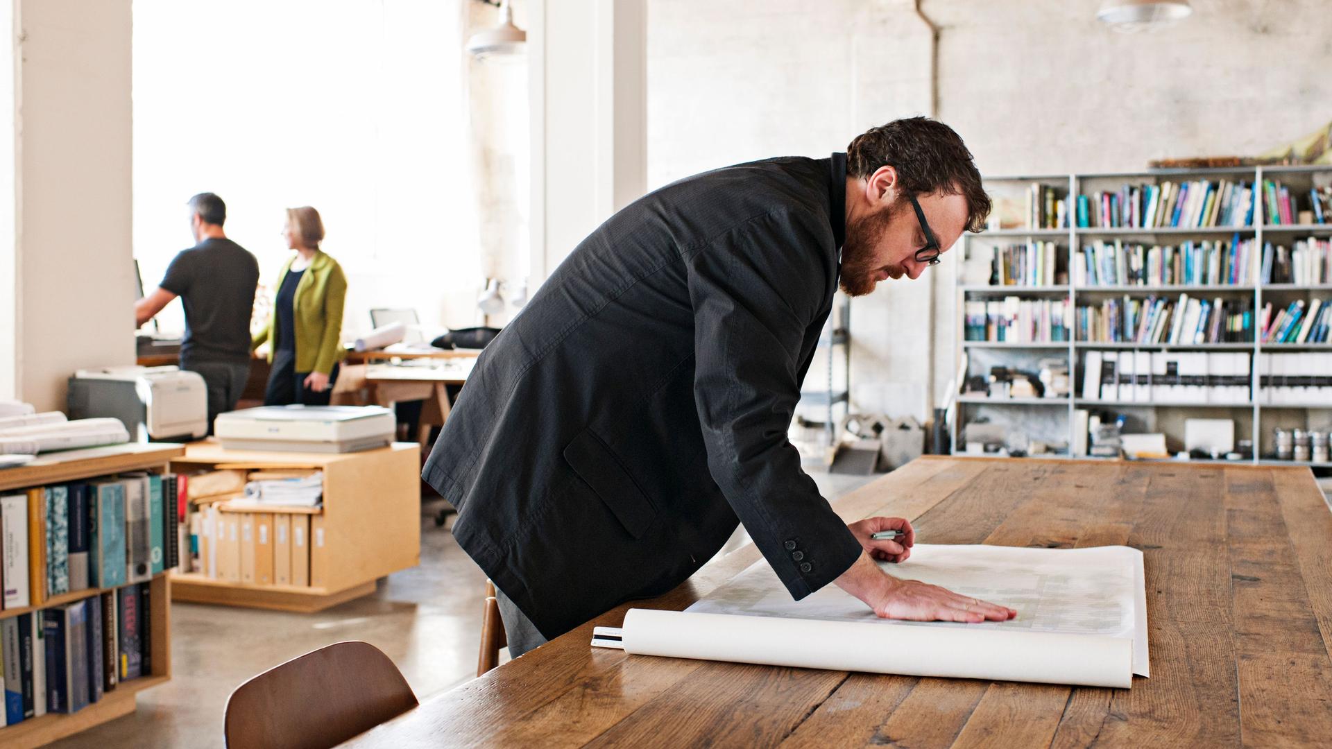 Ein Mann im Sakko prüft architektonische Pläne auf Papier, die auf einem Holztisch in einem modernen Büro liegen.