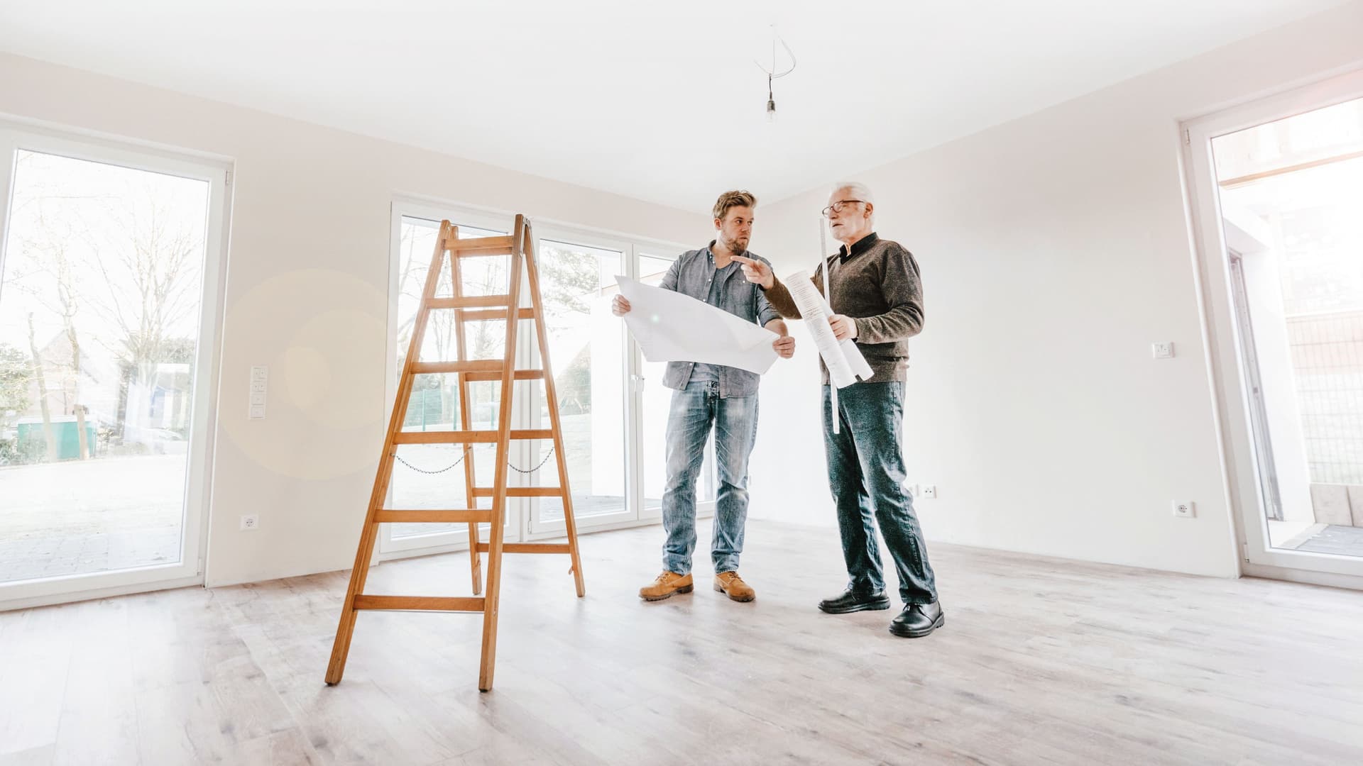 Ein Architekt prüft den Bauplan mit dem Eigentümer des neuen Hauses. Sie stehen dabei in einem leeren hellen Zimmer mit einer hölzernen Leiter.