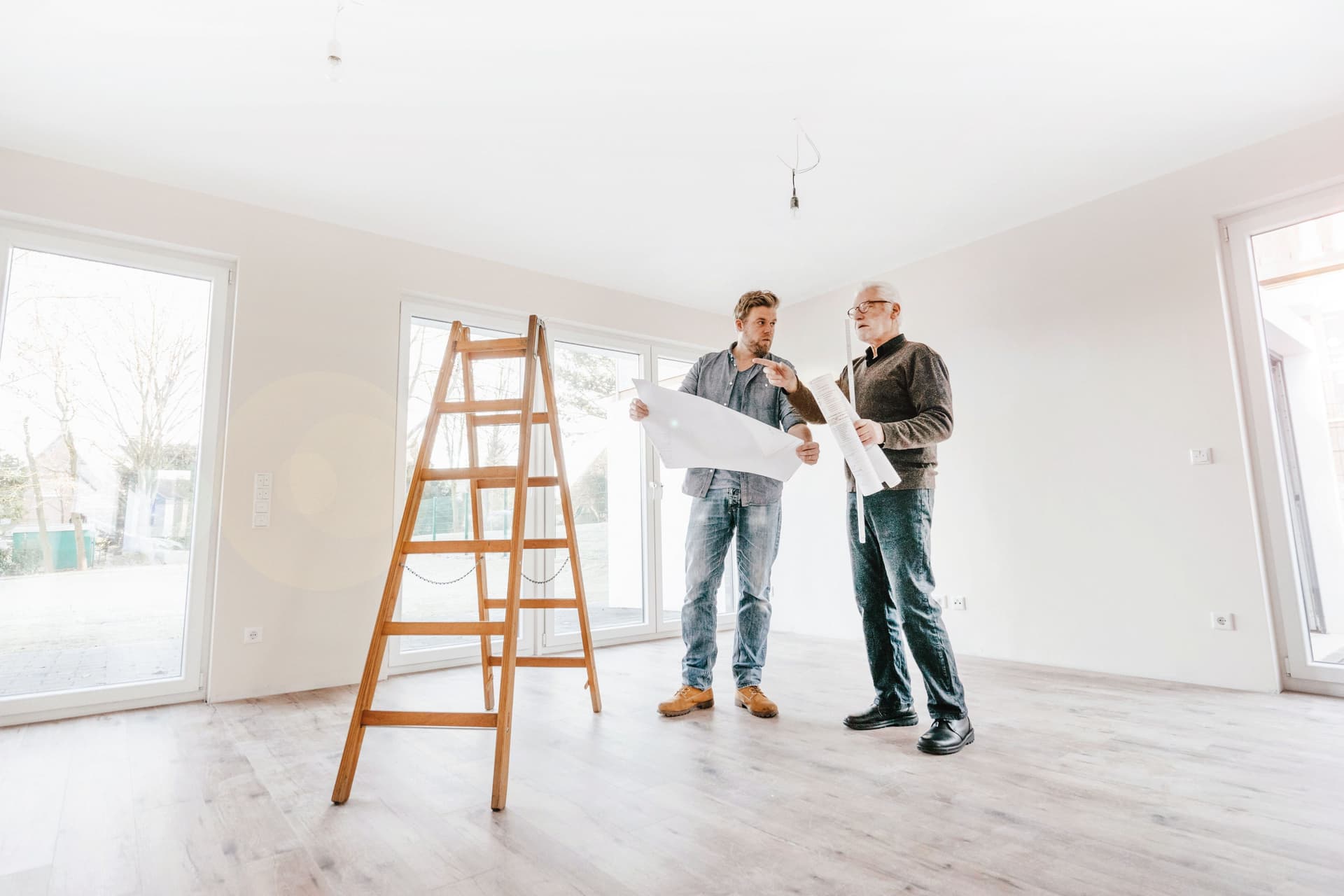 Ein Architekt prüft den Bauplan mit dem Eigentümer des neuen Hauses. Sie stehen dabei in einem leeren hellen Zimmer mit einer hölzernen Leiter.