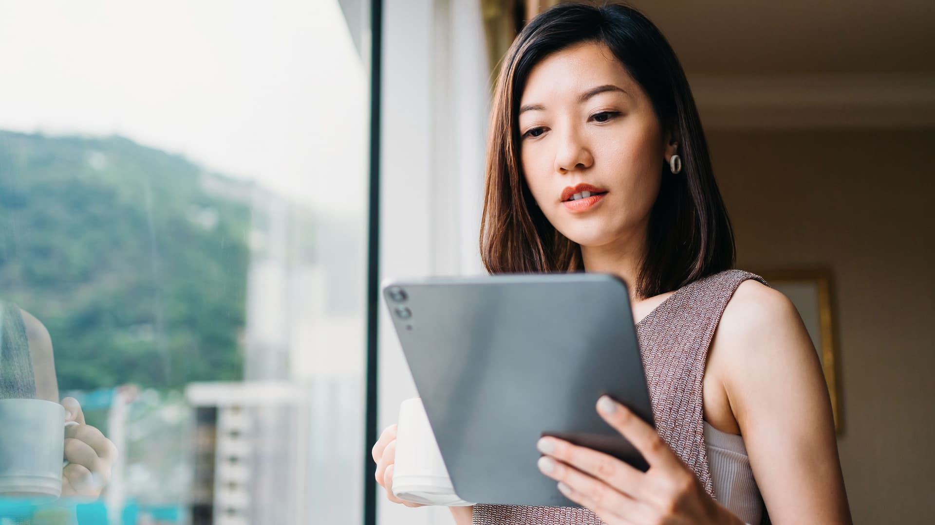 Frau trinkt Kaffee am Fenster während sie auf ihr Tablet schaut.