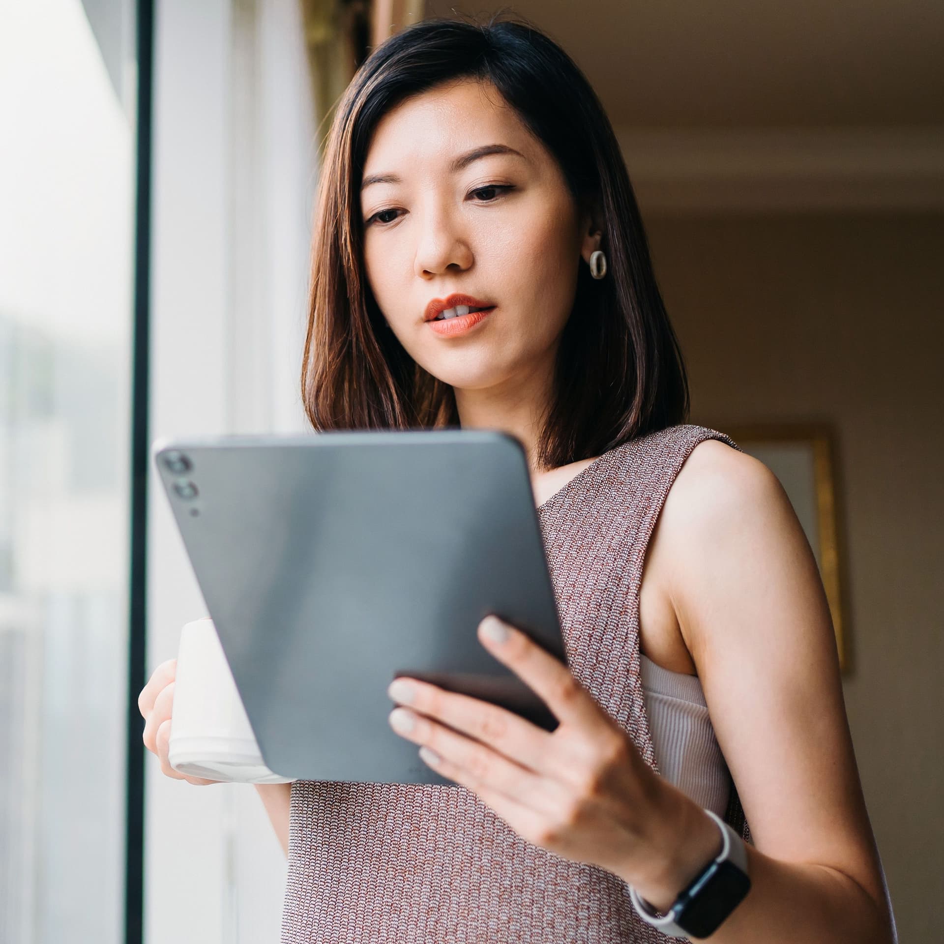 Frau trinkt Kaffee am Fenster während sie auf ihr Tablet schaut.