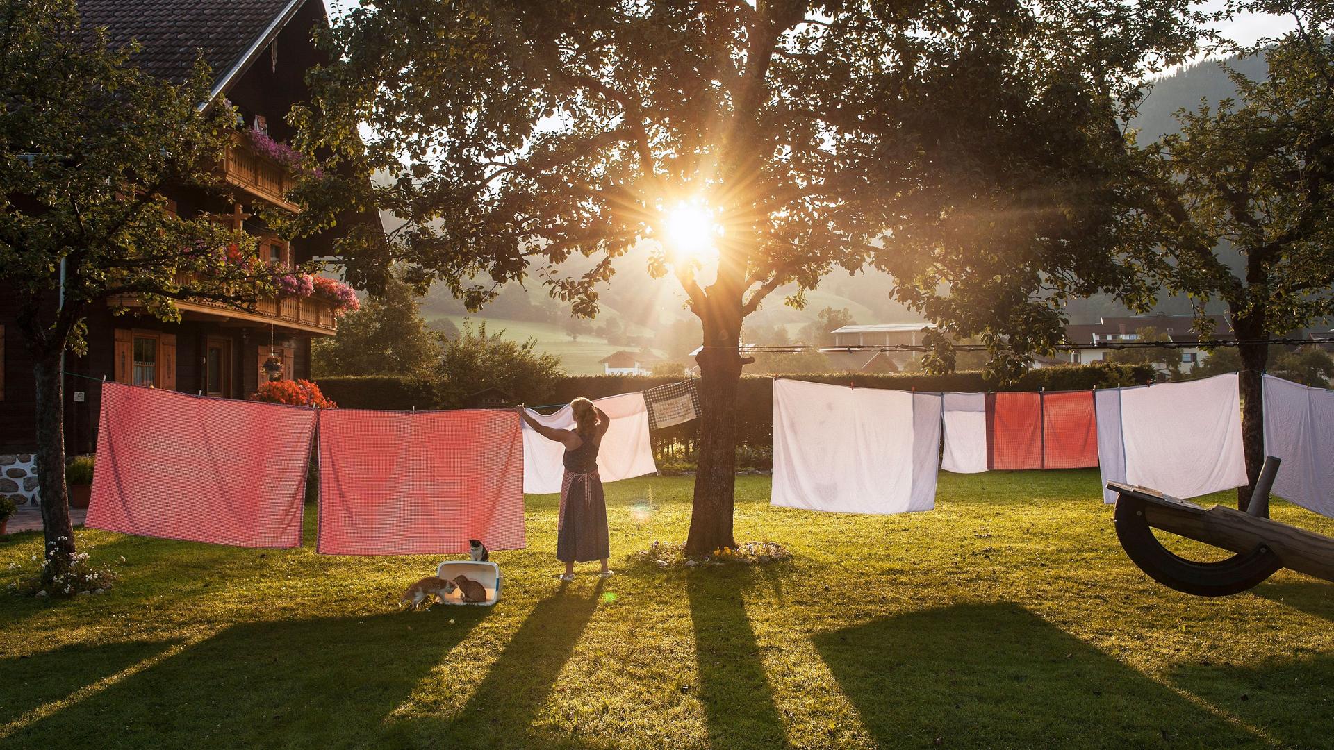 Ein Frau hängt in einem Garten Wäsche auf eine Wäscheleine. Im Hintergrund steht ein traditionelles österreichisches Haus. Die Sonne scheint und es ist warm.