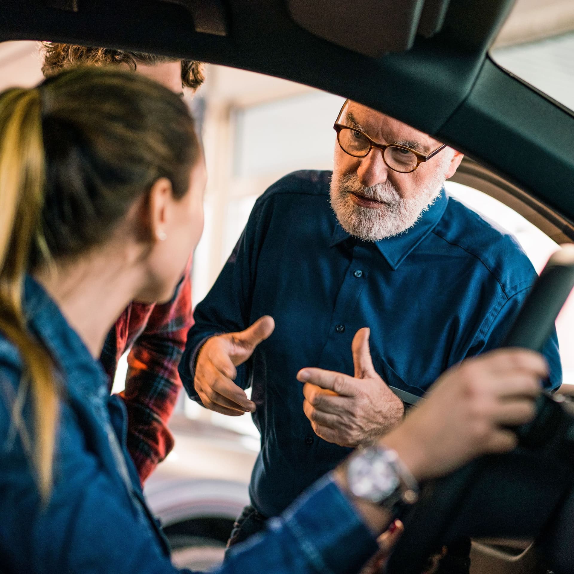 Ein Automechaniker spricht mit einer Frau, die am Steuer sitzt und erklärt ihr etwas.