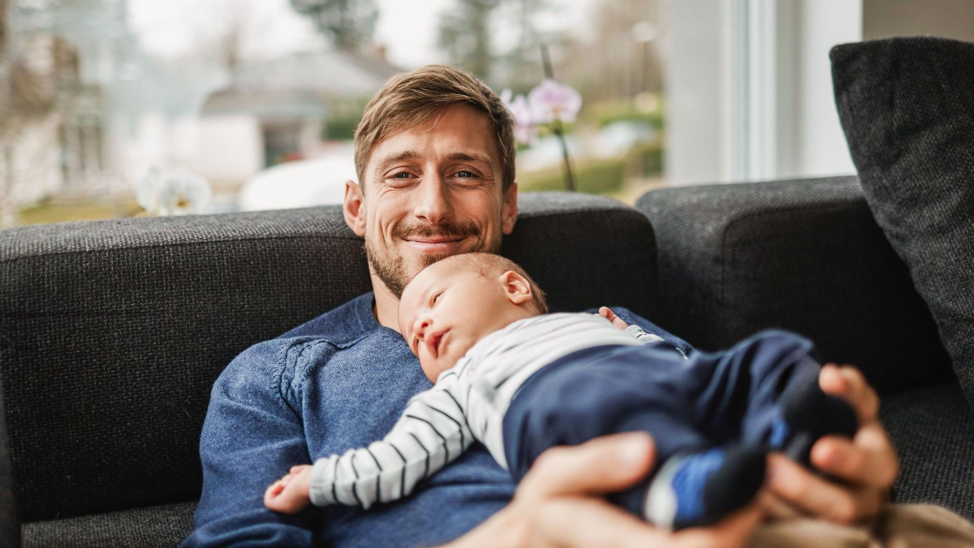 Ein lächelnder Vater liegt mit seinem Baby auf der Couch.