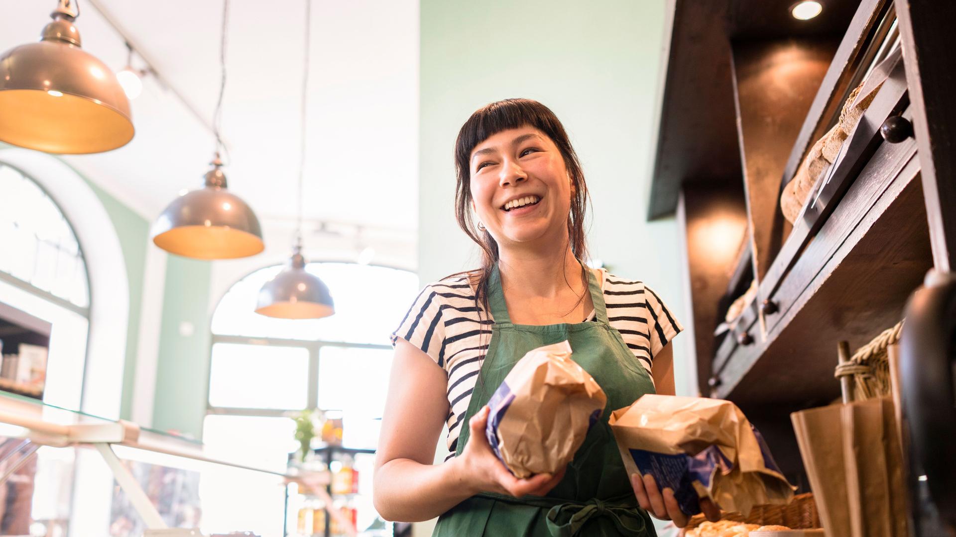 Lächelnde Besitzerin mit zwei eingepackten Broten in der Hand steht  zwischen einer Theke und Holzregalen in einer Bäckerei.