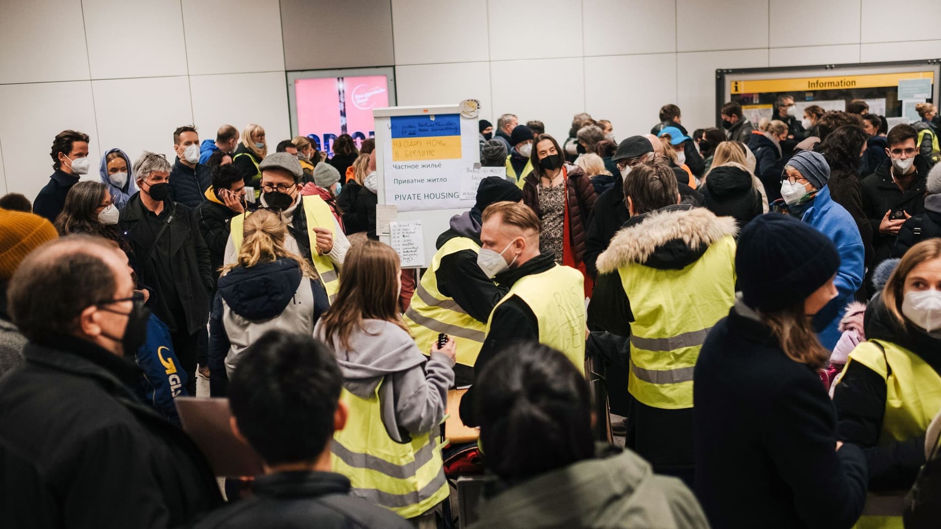 Zahlreiche Menschen auf einem deutschen Bahnhof stehen zusammen. Ukrainische Flüchtlinge werden von freiwilligen Helfern, die gelbe Warnwesten tragen, betreut.