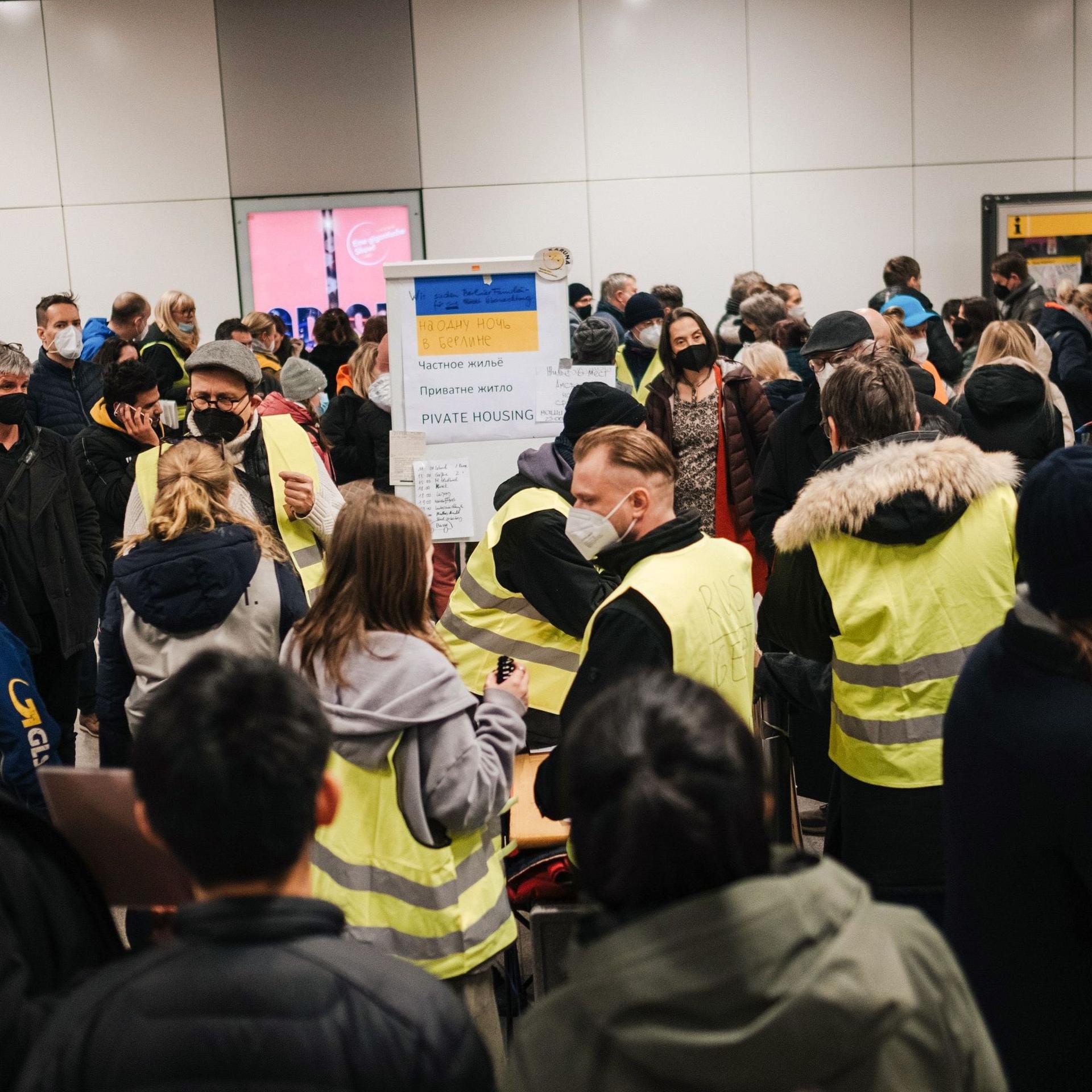 Zahlreiche Menschen auf einem deutschen Bahnhof stehen zusammen. Ukrainische Flüchtlinge werden von freiwilligen Helfern, die gelbe Warnwesten tragen, betreut.