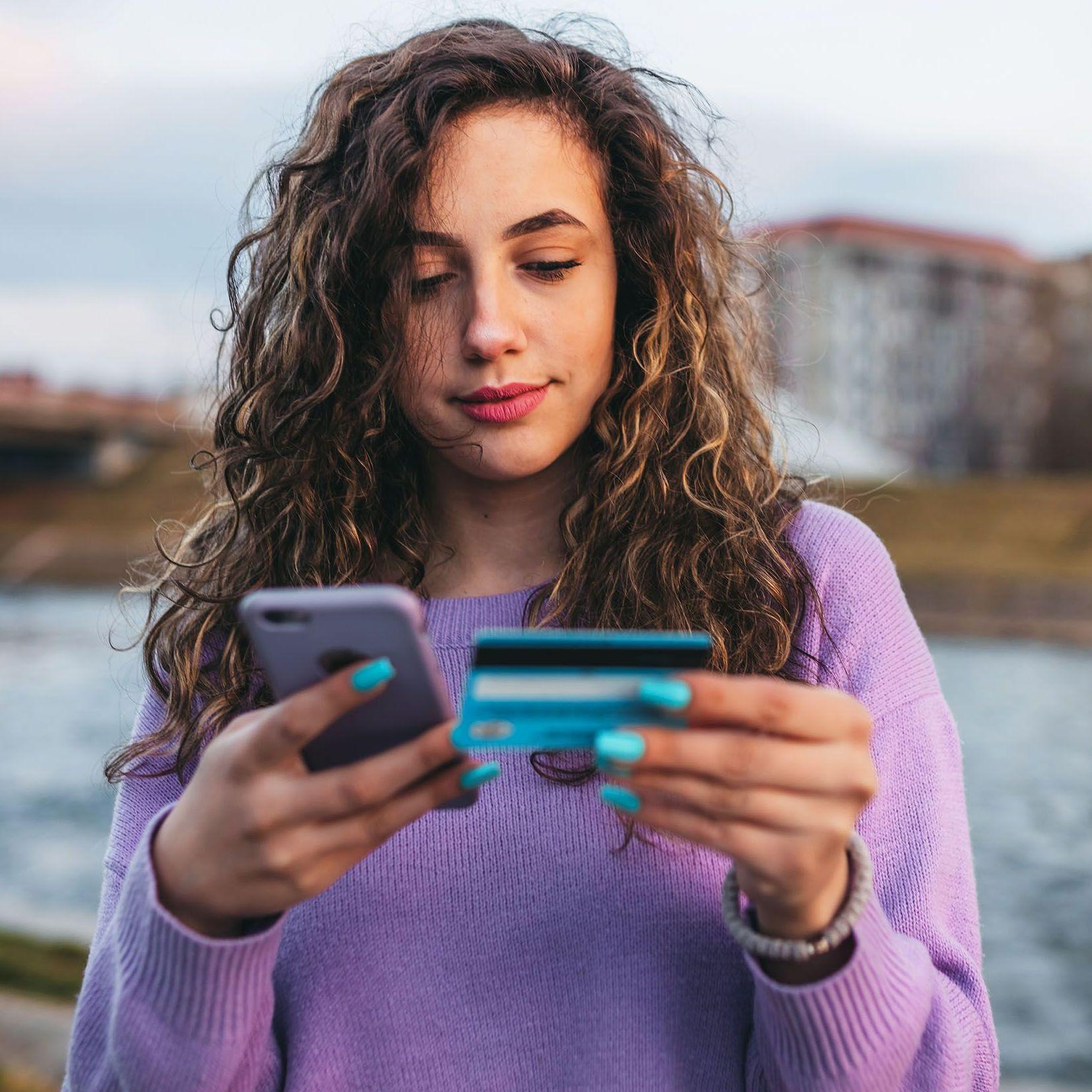 Eine junge Frau mit langen lockingen, braunen Haaren in einem violetten Pullover schaut auf ihr Smartphone und hält ihre Geldkarte in der Hand. 