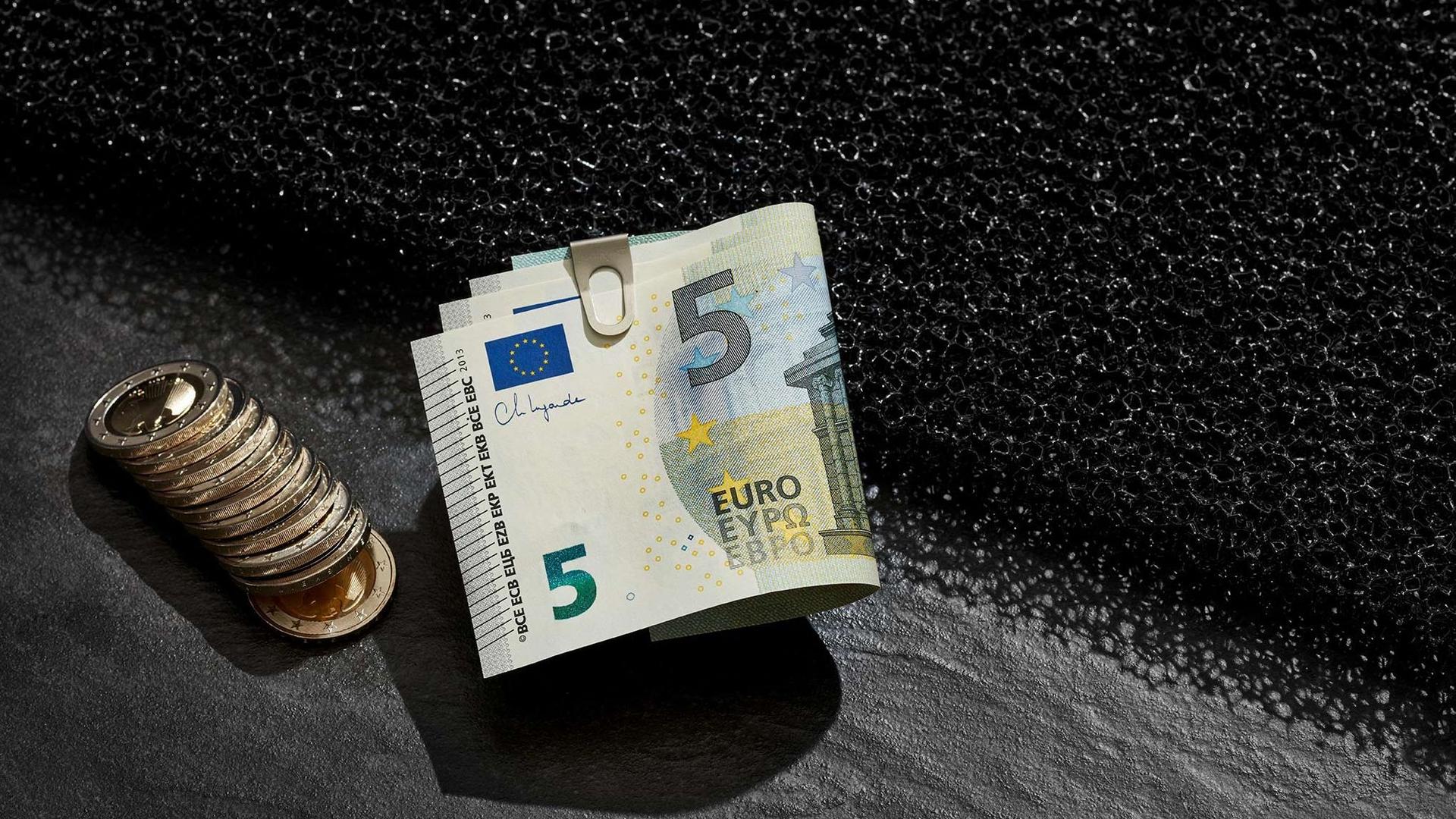 Inszeniertes Stillleben mit Eurogeldscheinen in einer Geldklammer und aufgereihten Münzen daneben. Sie liegen auf einem dunklen Untergrund.