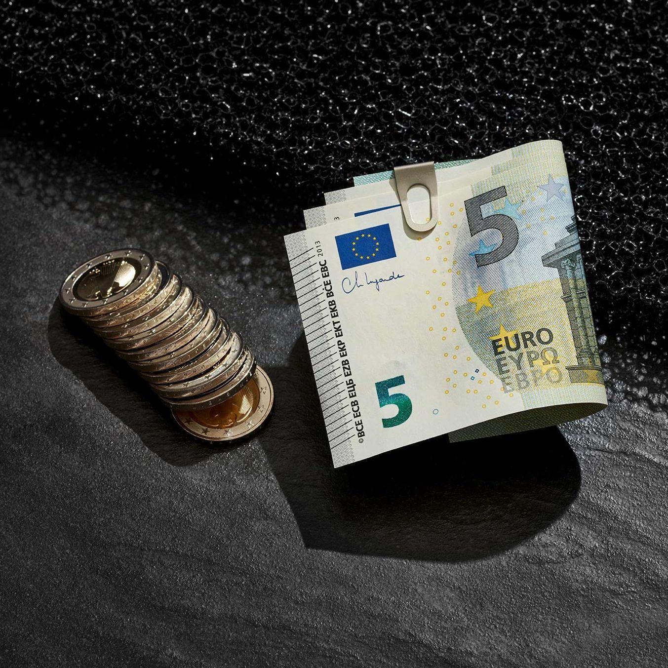 Inszeniertes Stillleben mit Eurogeldscheinen in einer Geldklammer und aufgereihten Münzen daneben. Sie liegen auf einem dunklen Untergrund.
