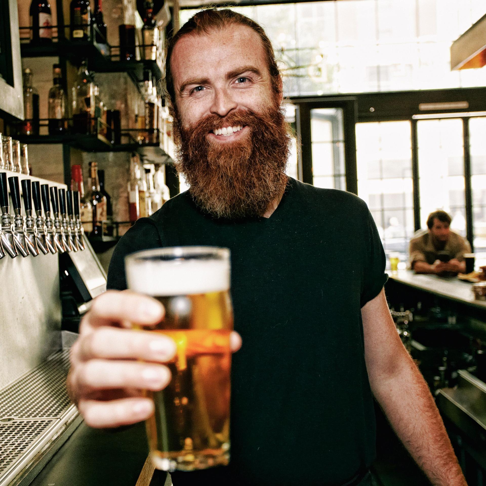 Ein junger Mann mit einem langen rötlichen Bart steht hinter einer Theke und streckt dem Betrachter lächelnd ein gefülltes Bierglas entgegen.