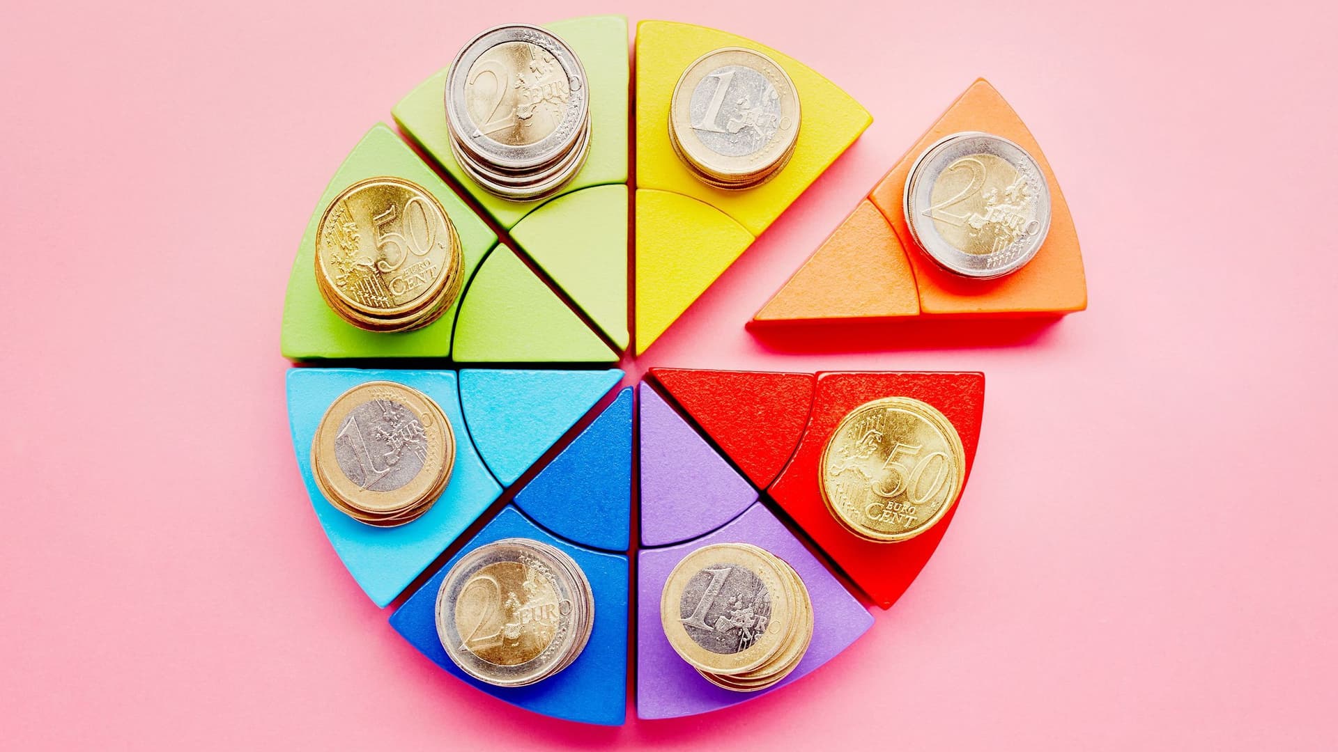 Ein Kuchendiagramm aus Bauklötzen und Münzen auf einem rosa Hintergrund.