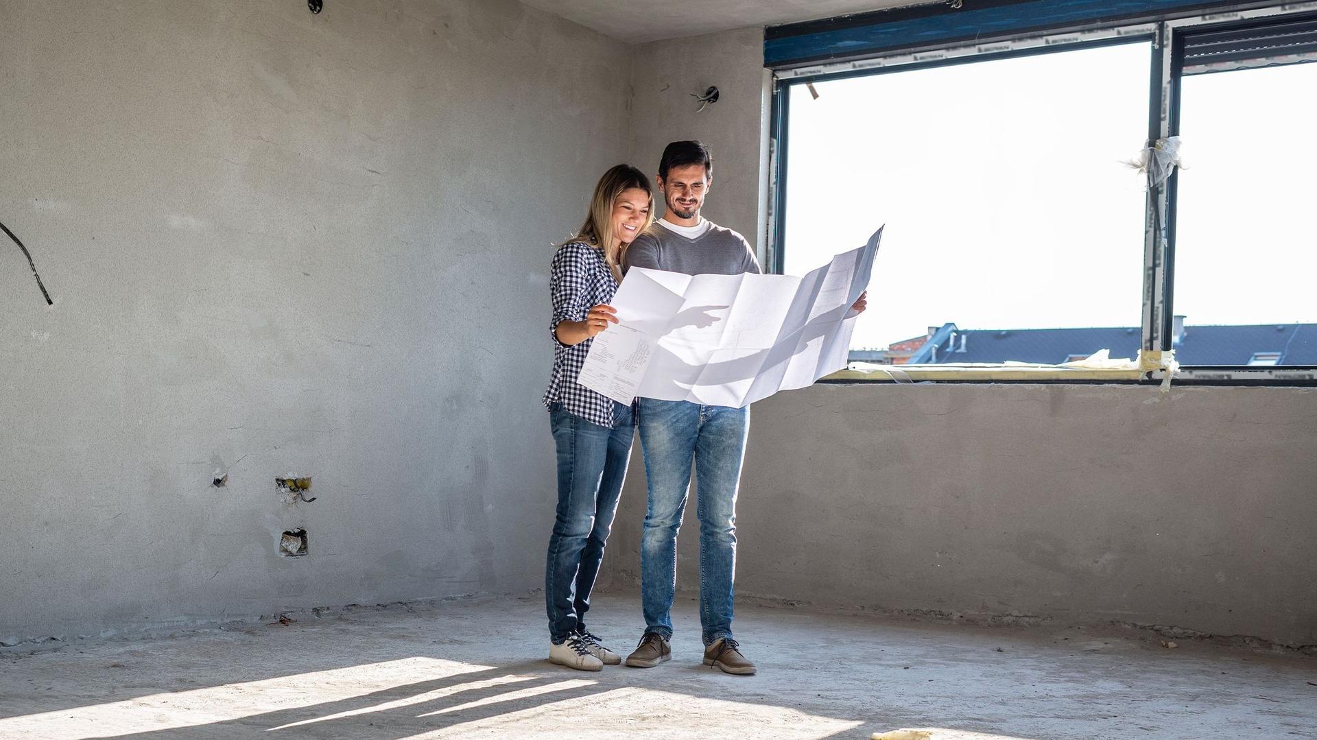 Eine Frau und ein Mann stehen inmitten eines Rohbaus auf einer Baustelle und sehen sich zusammen den Bauplan an. 