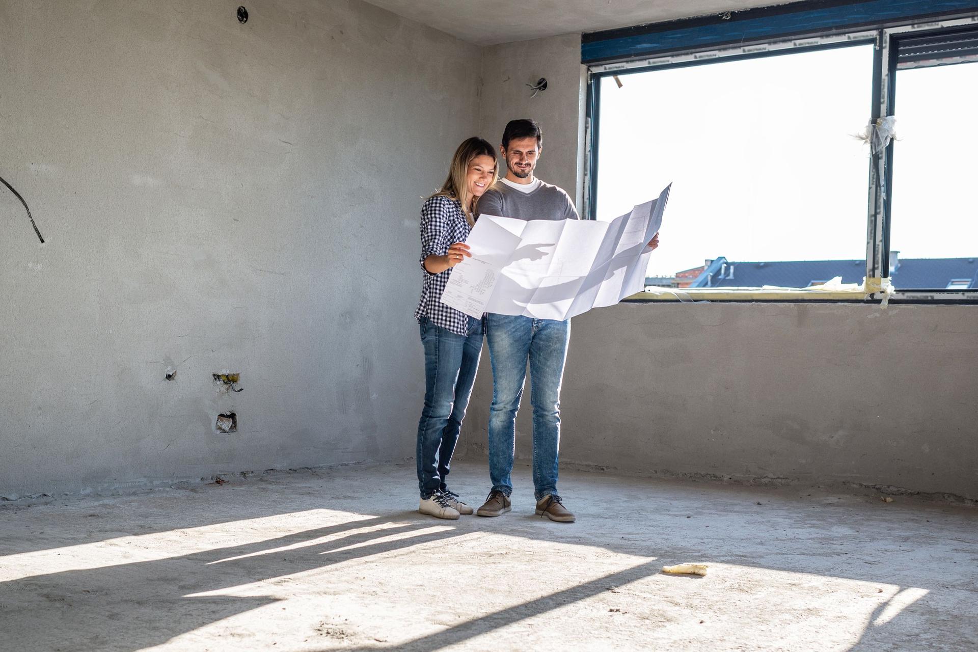Eine Frau und ein Mann stehen inmitten eines Rohbaus auf einer Baustelle und sehen sich zusammen den Bauplan an. 