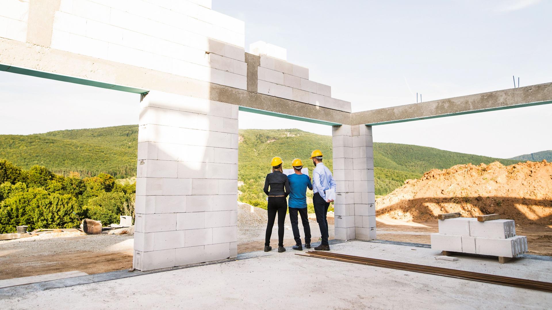 Drei Architekten und Arbeiter mit gelben Helmen und einem Plan in der Hand auf der Baustelle eines Rohbaus.