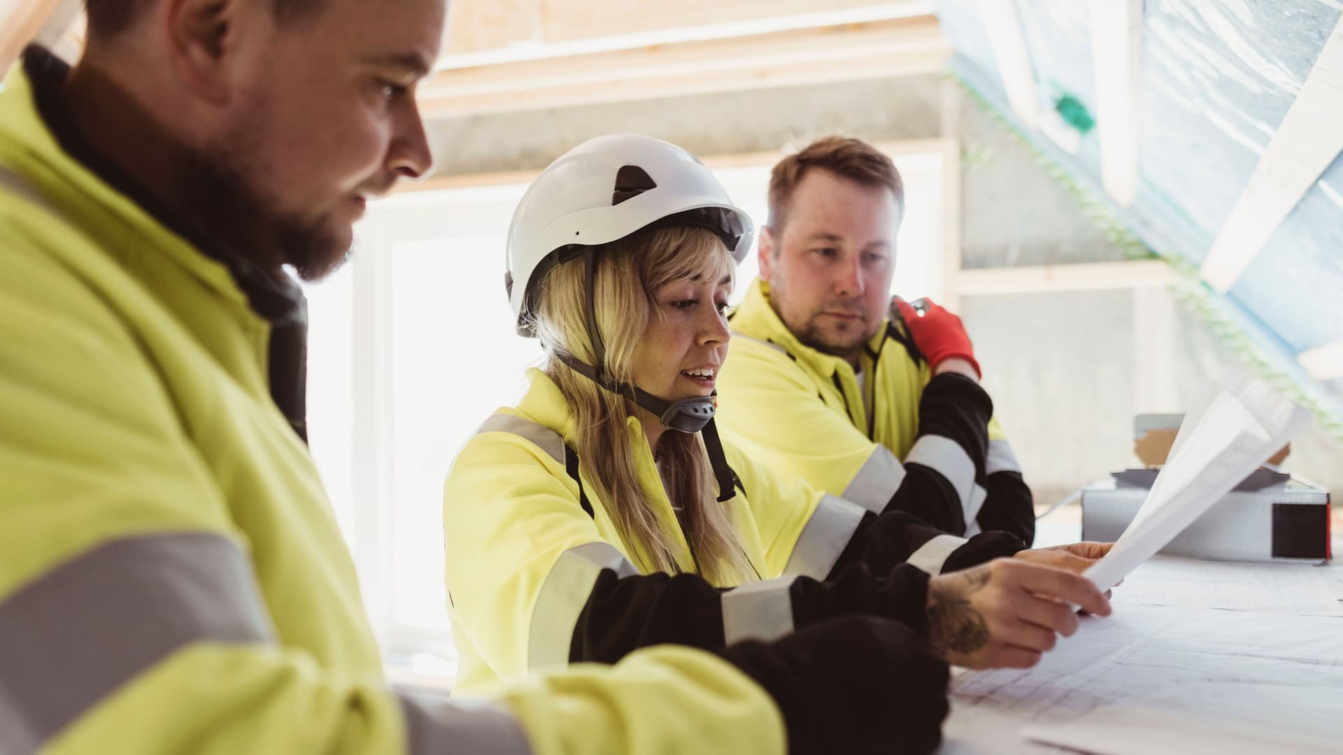 Eine Frau mit Helm und Arbeitsjacke diskutiert über Grundriss auf einem Papier mit zwei Männern in reflektierender Arbeitsjacke.