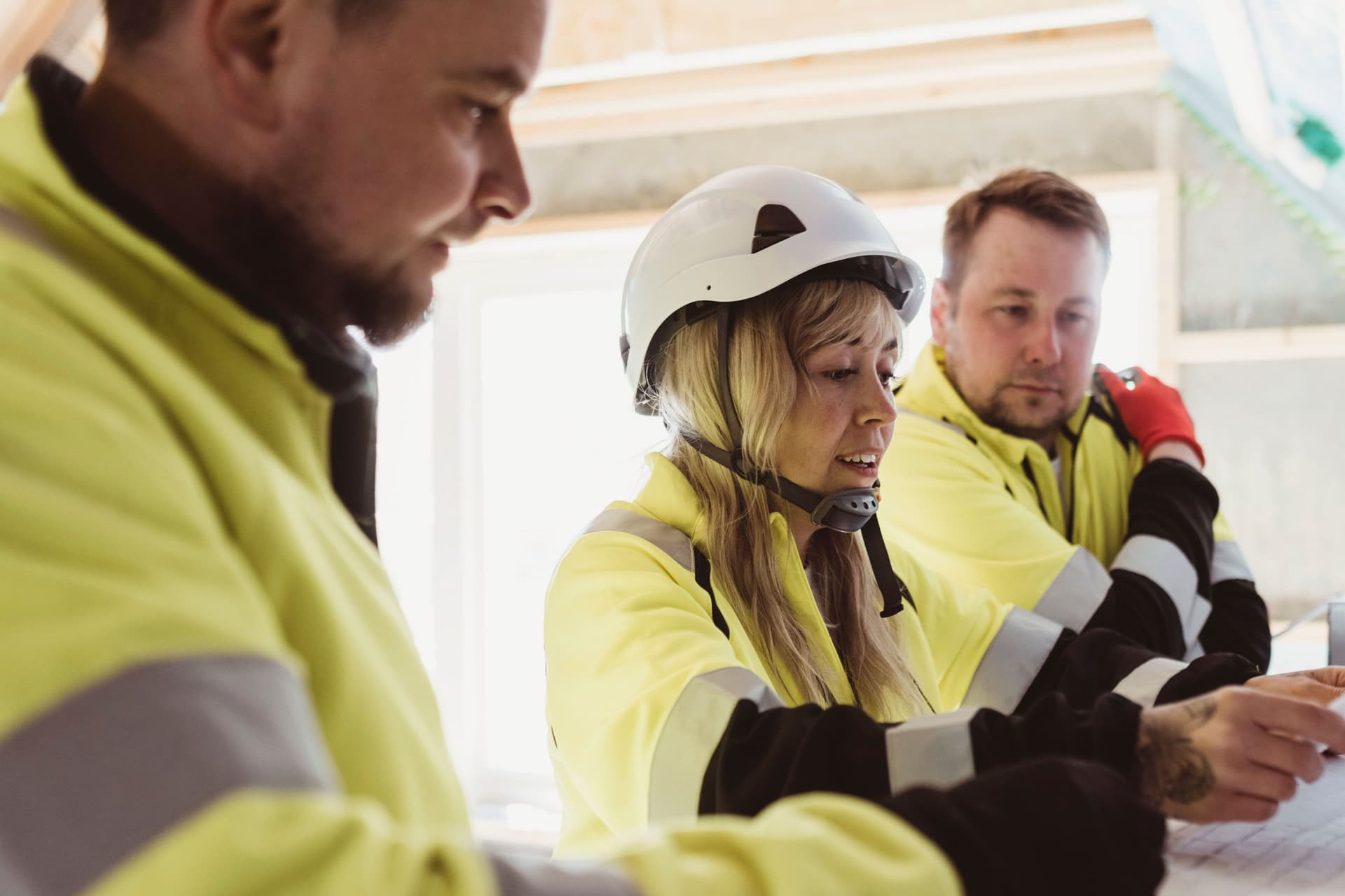 Eine Frau mit Helm und Arbeitsjacke diskutiert über Grundriss auf einem Papier mit zwei Männern in reflektierender Arbeitsjacke.