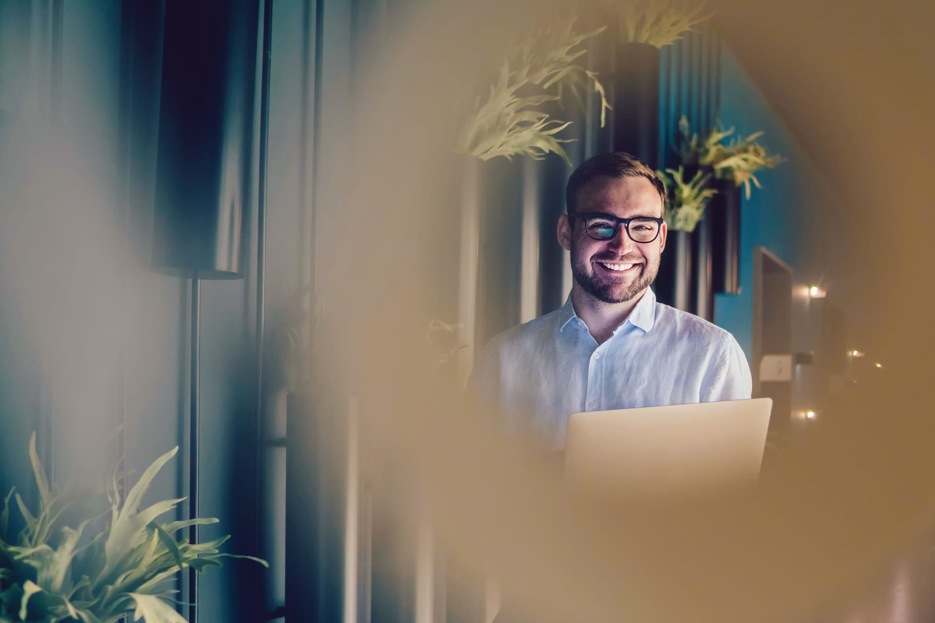 Blick durch die Unschärfe auf einen lachenden jungen Mann mit Laptop in der Hand. Er steht in einem Gang eines Bürogebäudes.