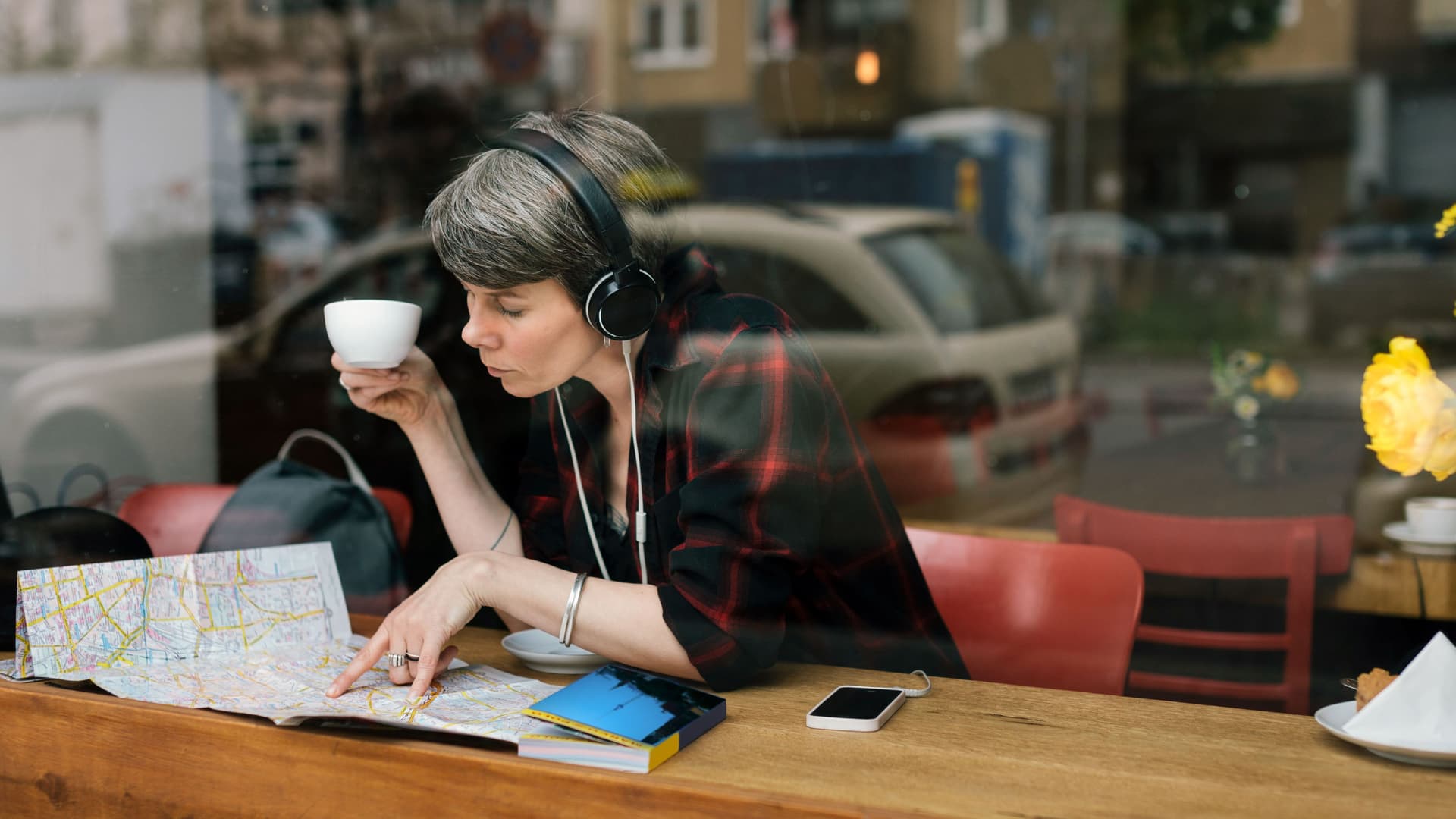 Blick durch ein Caféfenster auf eine Frau mittleren Alters, die eine Tasse in der Hand hält und Kopfhörer auf hat. Sie blickt auf einen Stadtplan.