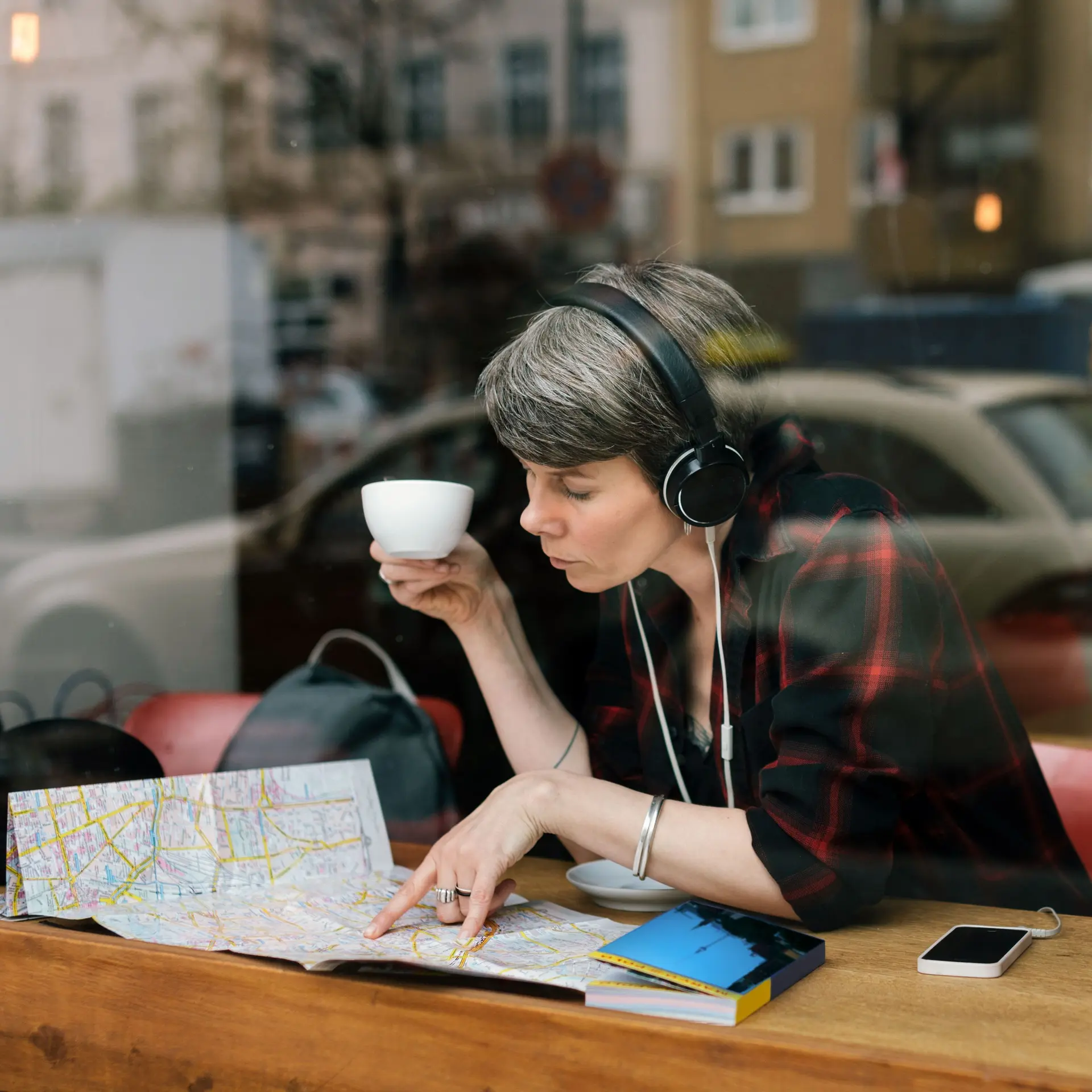 Blick durch ein Caféfenster auf eine Frau mittleren Alters, die eine Tasse in der Hand hält und Kopfhörer auf hat. Sie blickt auf einen Stadtplan.