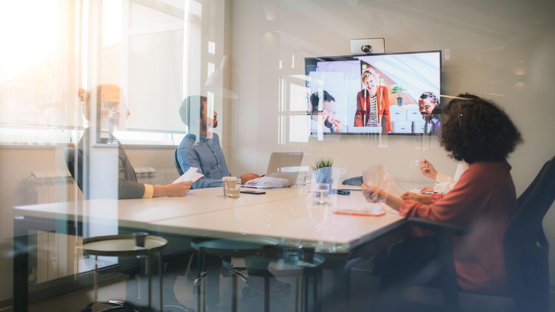 Blick durch Glasscheiben in einen Meetingraum mit Videoprojektion. In dem Meetingraum sitzen vier Personen.