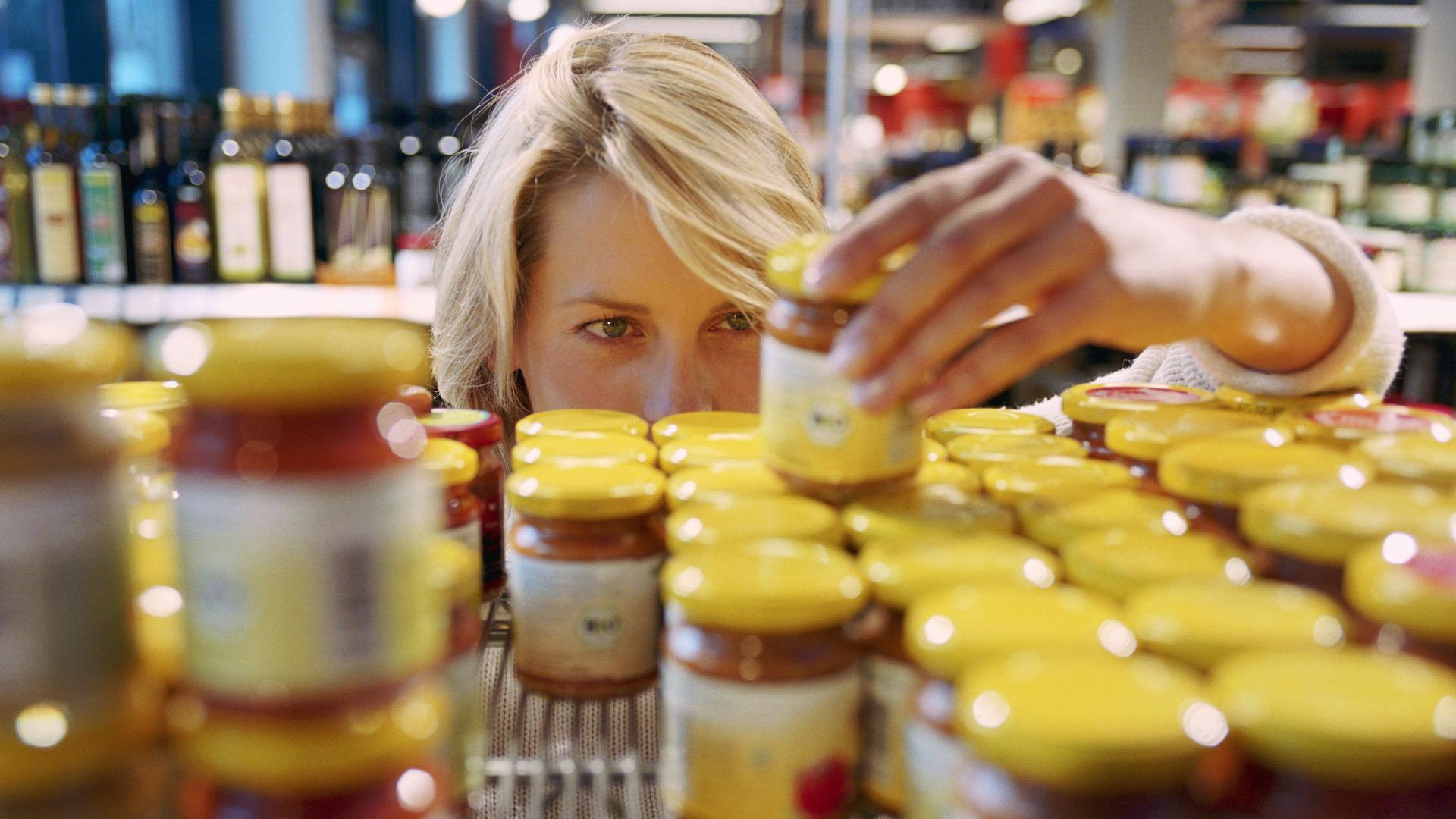 Blick durch ein Supermarktregal mit Gläschen mit gelben Deckeln. Man sieht den Kopf einer Frau, die sich ein Gläschen anguckt was sie in der Hand hält.