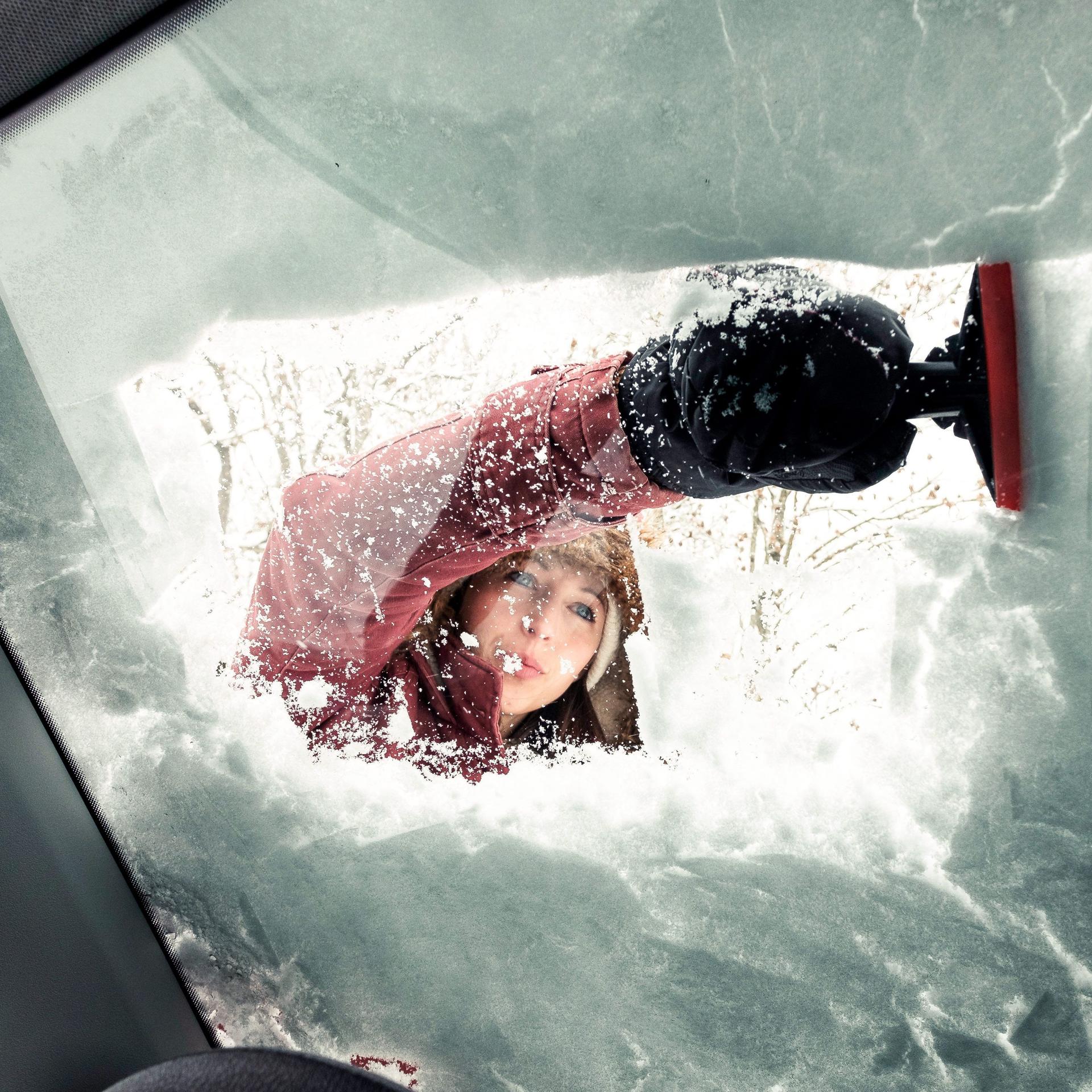 Blick vom Innenraum eines Autos durch eine Autoscheibe, von der eine junge Frau Schnee kratzt.
