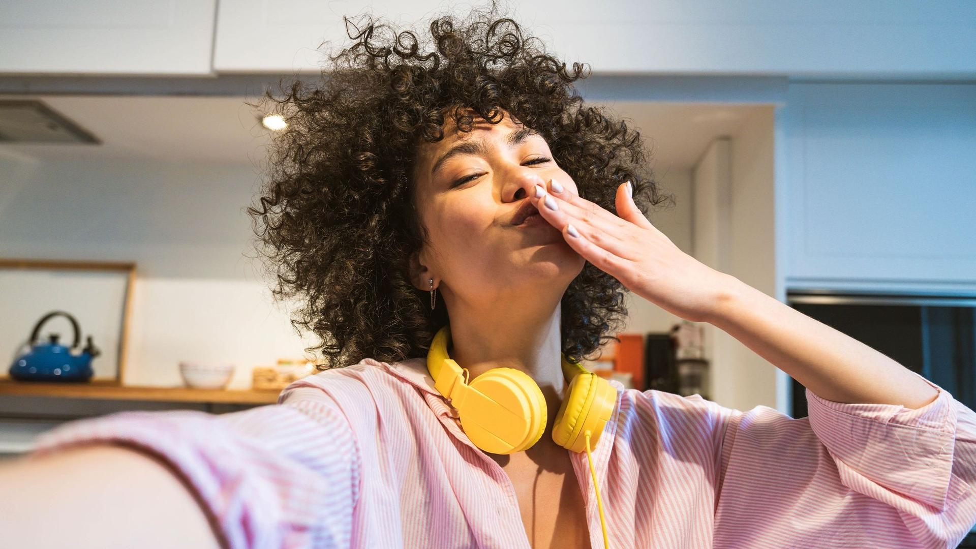 Eine junge, glückliche Frau mit gelben Kopfhörern um den Hals macht ein Selfie mit Luftkuss in ihrer neuen Wohnküche.
