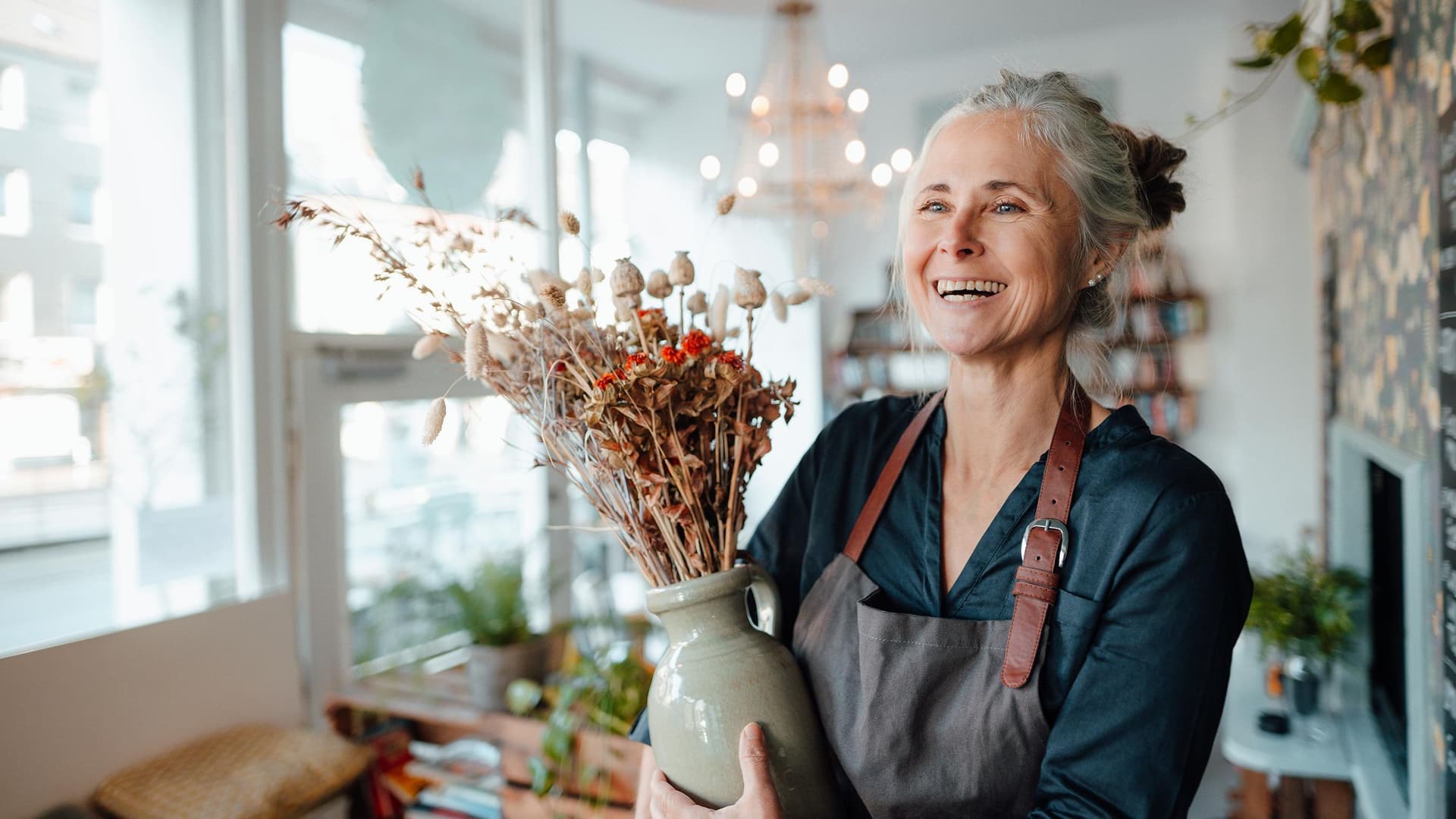Eine ältere Frau mit Schürze und grauen Haaren steht in einem schönen und lichtdurchfluteten Blumenladen. Sie lacht ausgelassen und hat eine Vase mit einem Blumengesteck in der Hand.