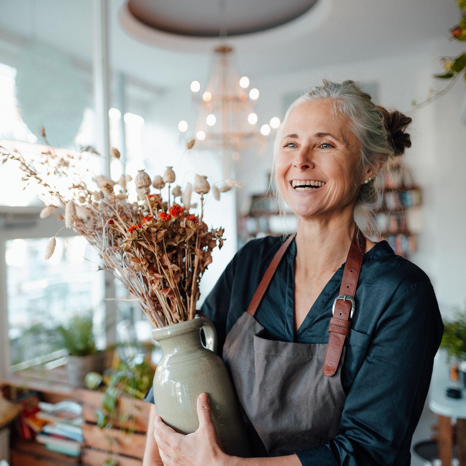 Eine ältere Frau mit Schürze und grauen Haaren steht in einem schönen und lichtdurchfluteten Blumenladen. Sie lacht ausgelassen und hat eine Vase mit einem Blumengesteck in der Hand.