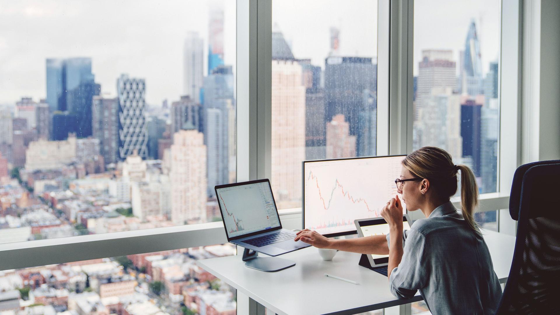 Konzentriert arbeitende Frau vor einem Panoramafenster sitzt an einem Schreibtisch mit zwei Bildschirmen. Im Hintergrund ist eine verschwommene Skyline zu erkennen.