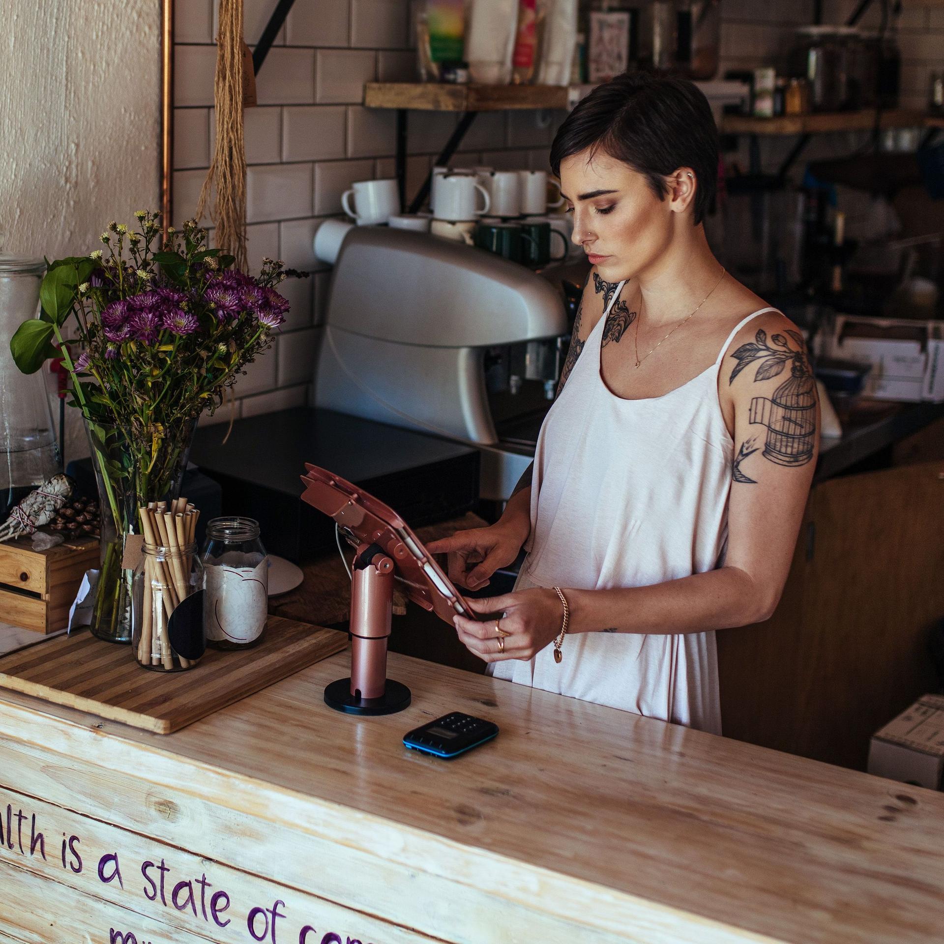 Junge Frau mit kurzen Haaren steht mit einem Tablet hinter dem Holz-Theresen in einem Café.