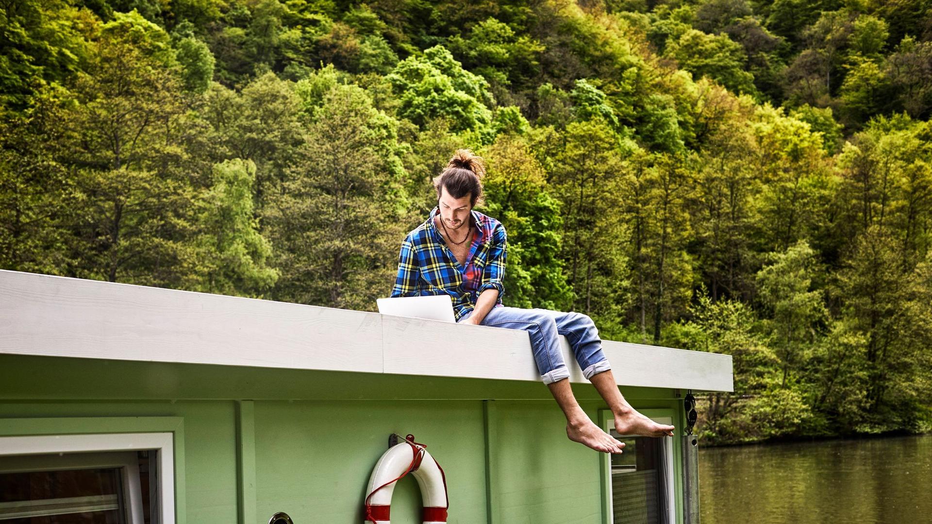 Ein junger Mann sitzt barfuß auf dem Dach eines Hausboots, auf einem von grünen Laubbäumen umgebenen See, und schaut auf seinen Laptop.