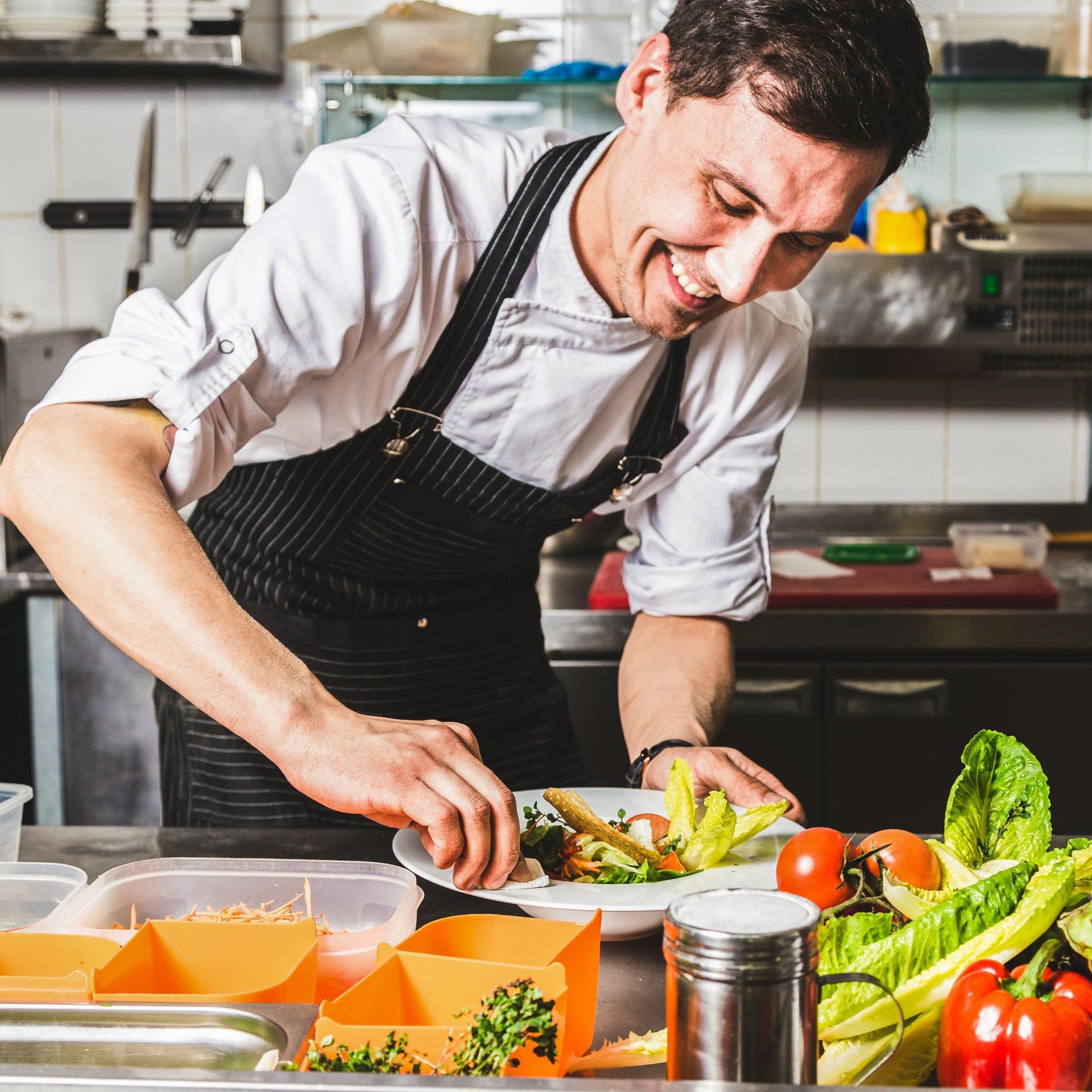 Professioneller Koch richtet lächelnd einen Salat in einer Restaurantküche an.