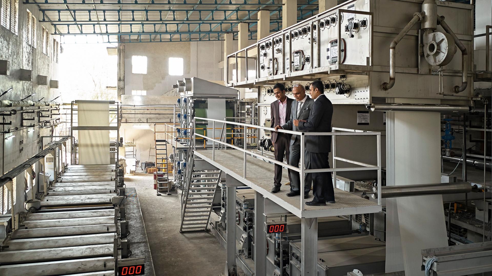 Drei Männer im Anzug, die auf einer erhöhten Plattform steht und das Fabriklayout in der Färberei und Druckerei in Mumbai studieren.
